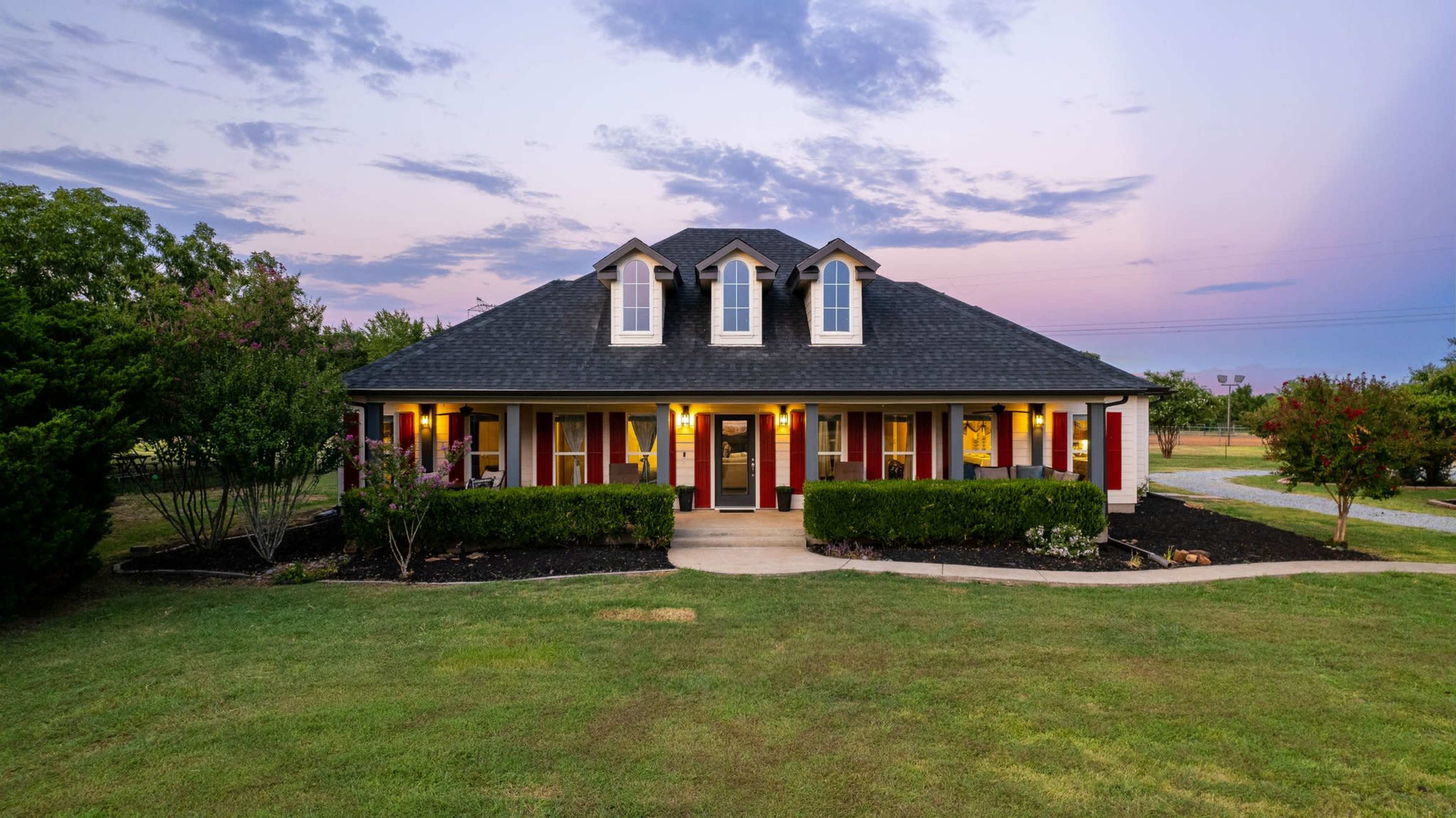 A two-story house with a dark roof and red shutters is surrounded by manicured lawns and shrubs under a twilight sky.