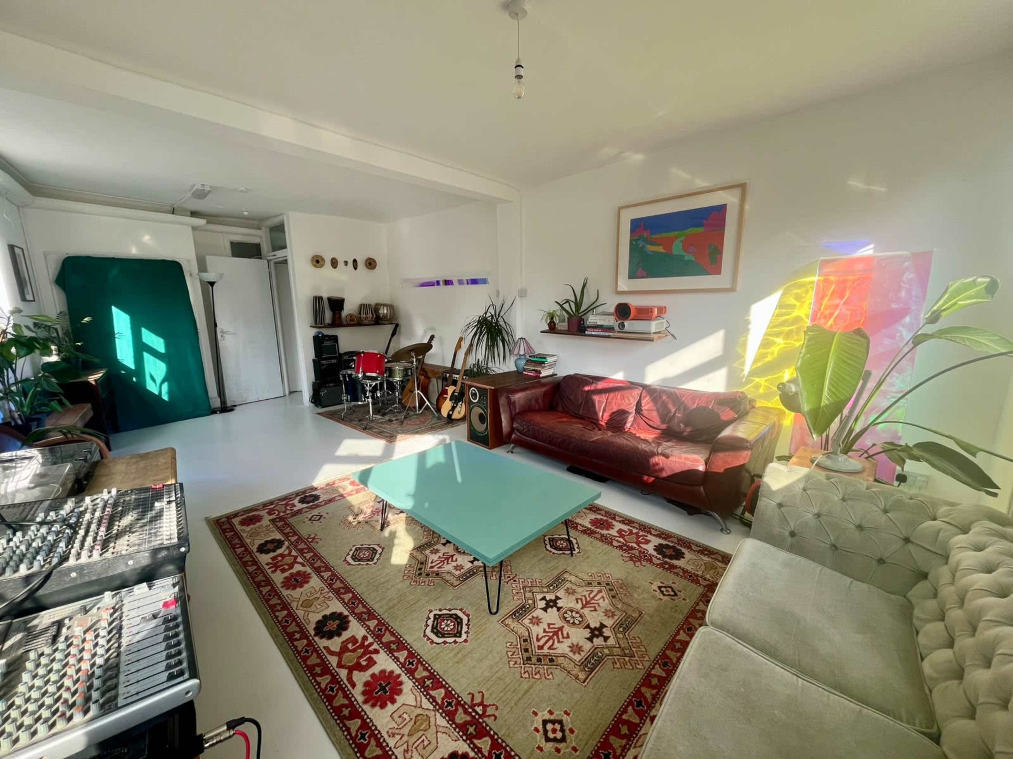 The room features a cozy living area with a red sofa, a light green coffee table, and a patterned rug in front of a variety of plants and colorful artwork on the walls.