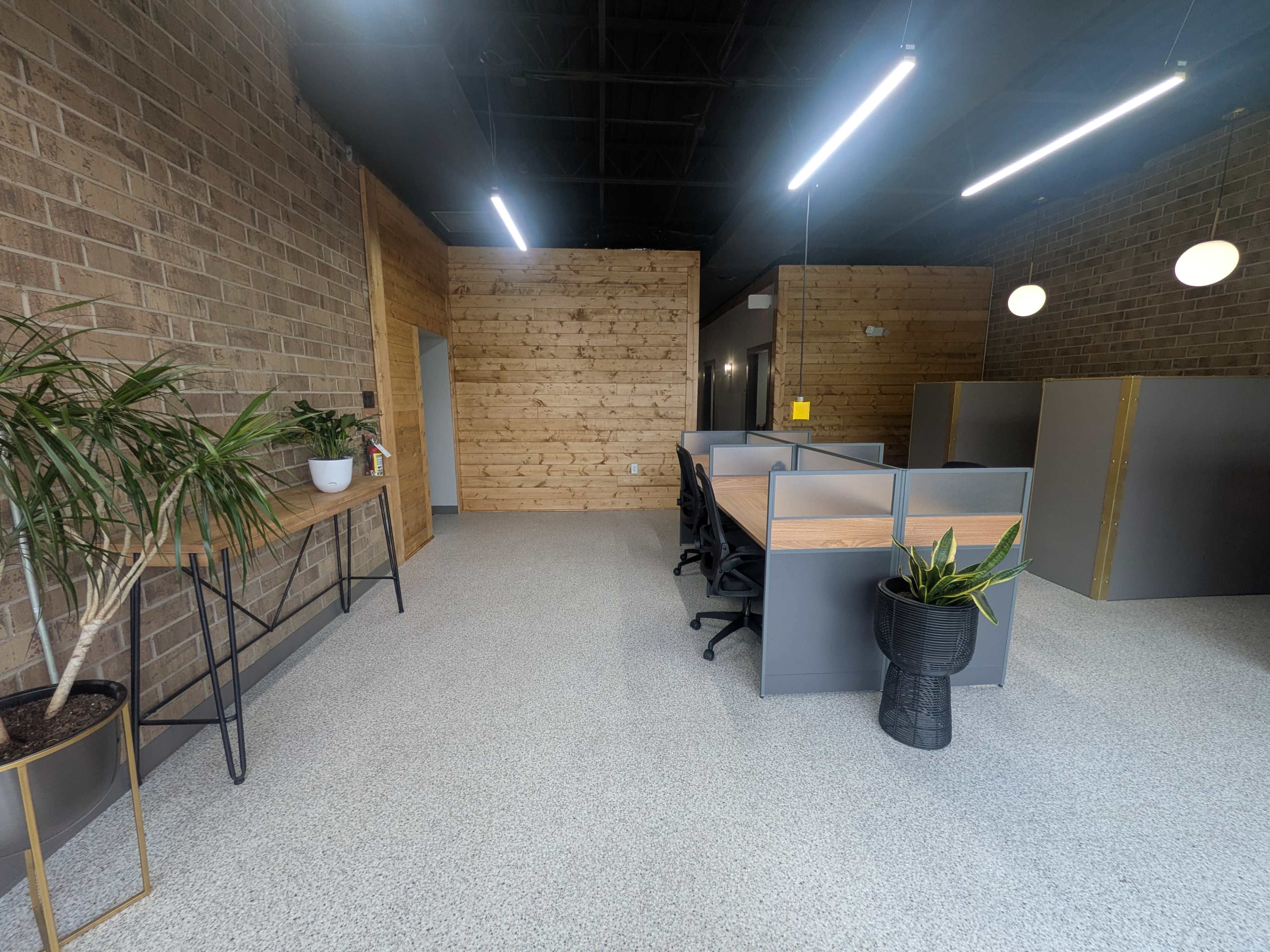 The image shows a modern office space with exposed brick walls, wooden paneling, and several workstations separated by dividers.