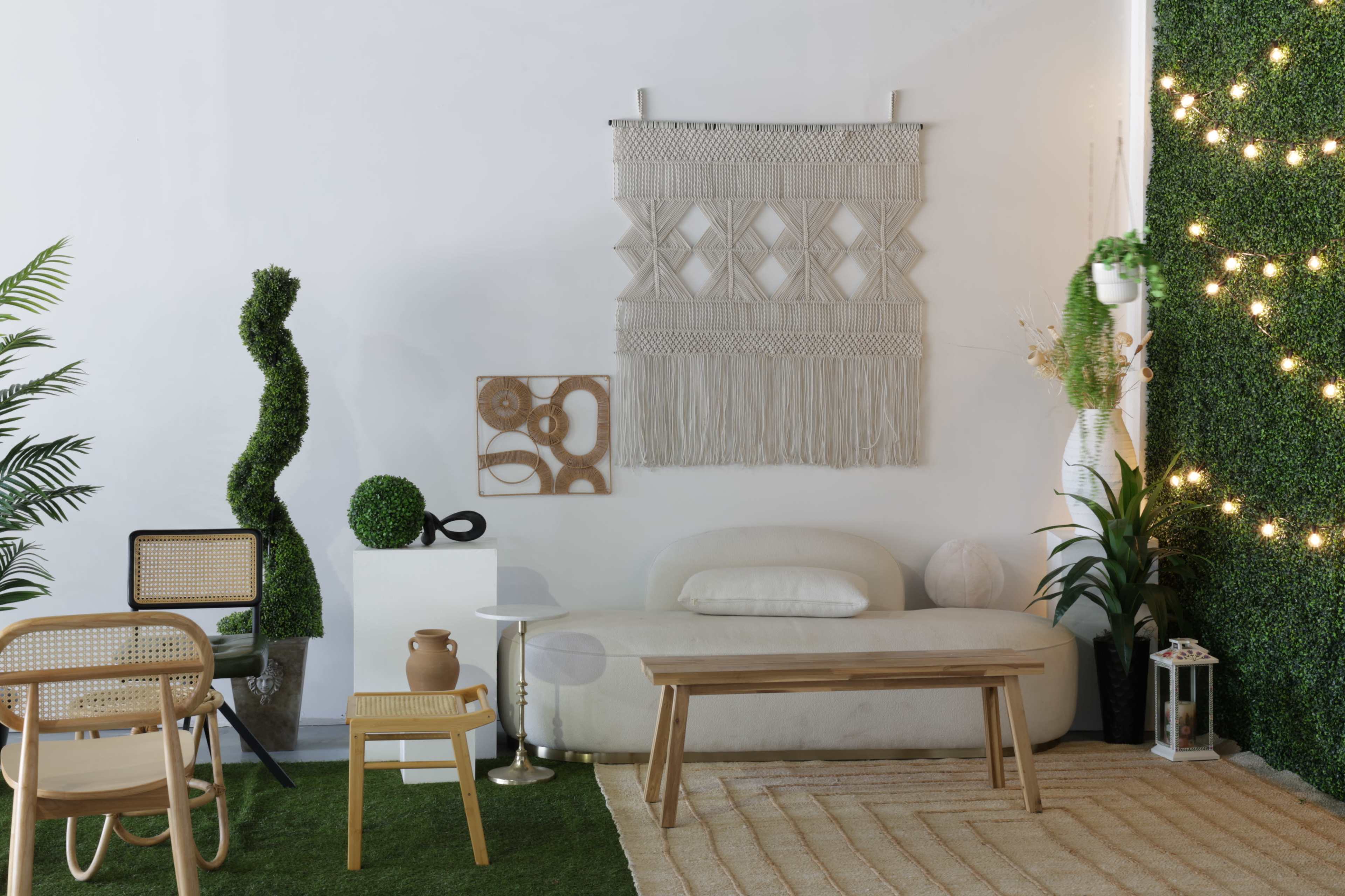 The image shows a modern living room with a white macramé wall hanging, a light-colored sofa, wooden furniture, and decorative greenery.