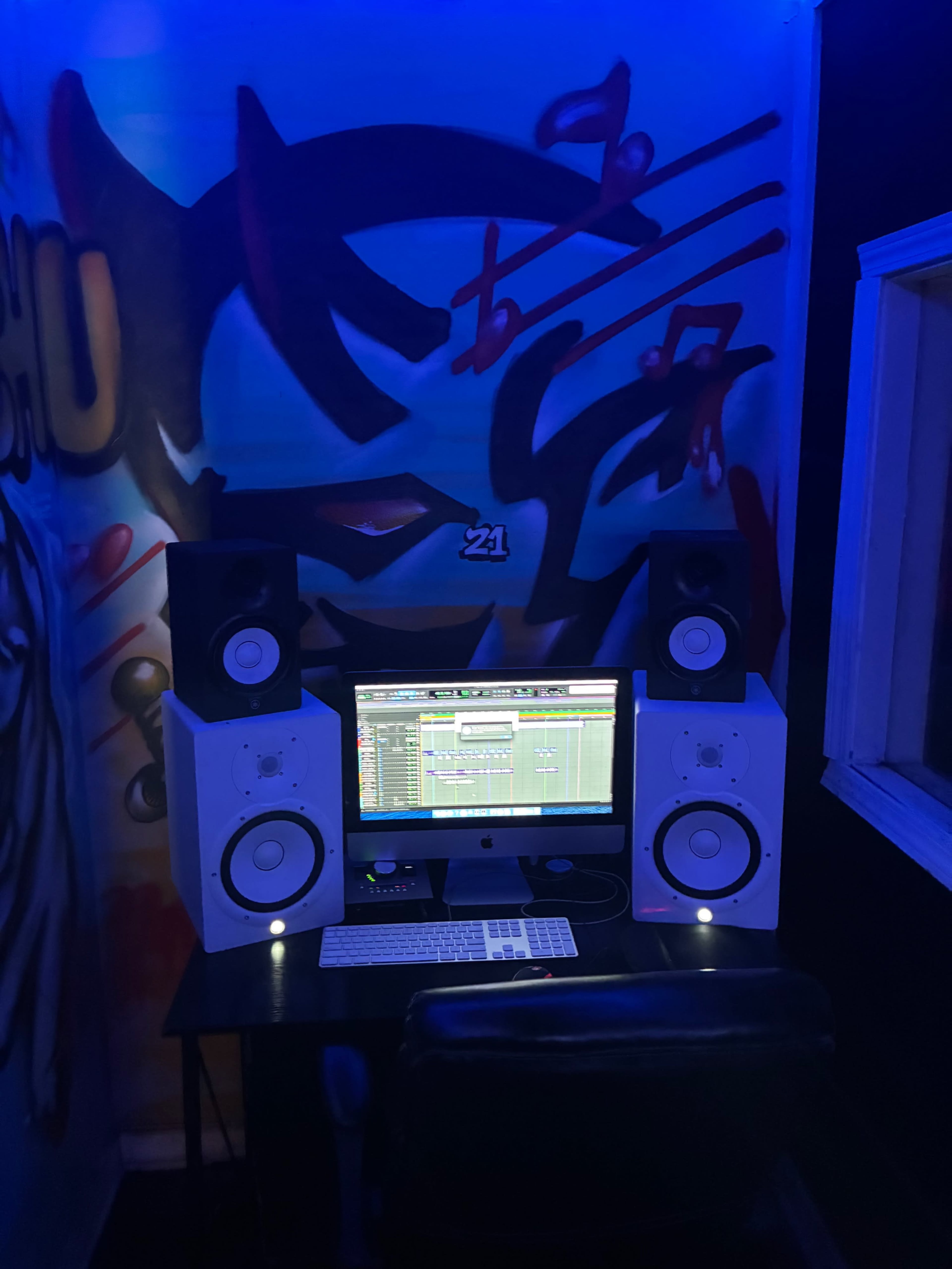 The image shows a music production setup with an Apple computer, speakers, and a chair, illuminated by blue light against a graffiti-adorned wall.