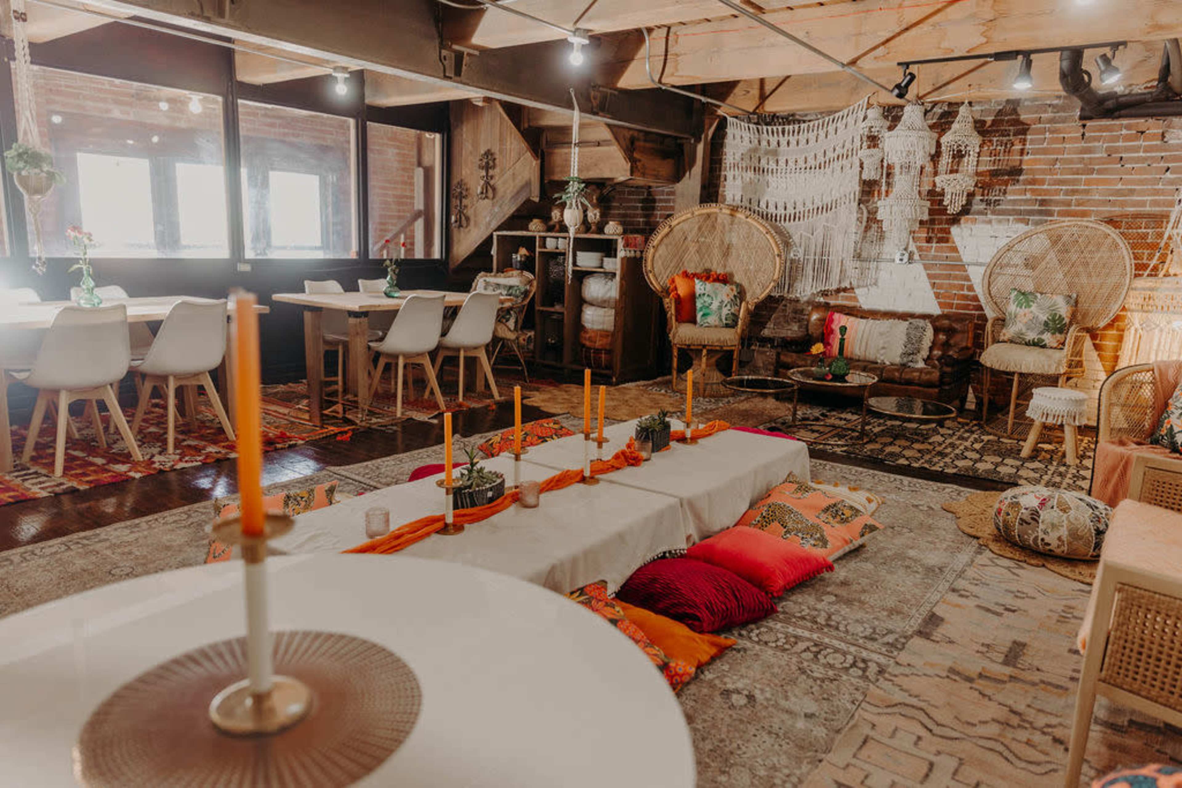 A cozy interior space featuring a mix of seating arrangements, including dining chairs and floor cushions, with decorative elements like hanging textiles and plants.