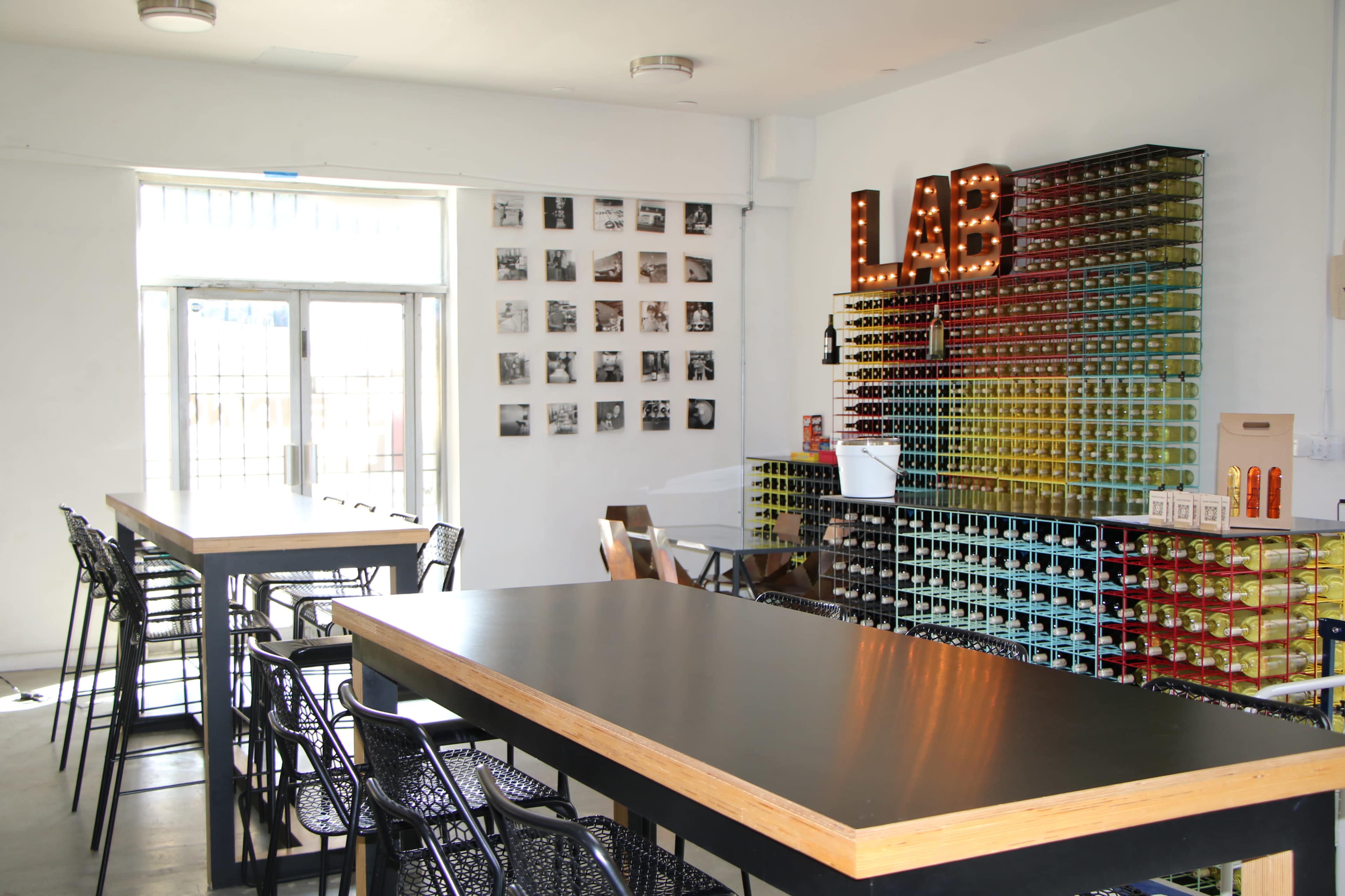 A modern café features a long black table, metal chairs, and a colorful wall of stacked containers, with a sign that reads "LAB" illuminated by bulbs.