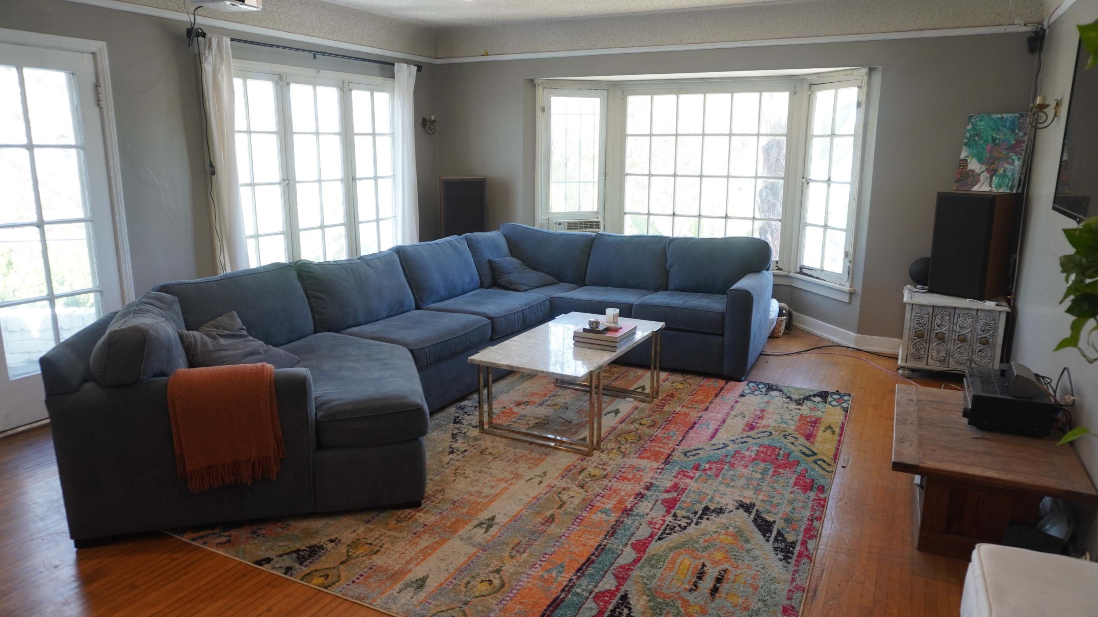 A spacious living room features a large blue sectional sofa, a glass coffee table, and colorful area rugs, with bright natural light streaming in through multiple windows.