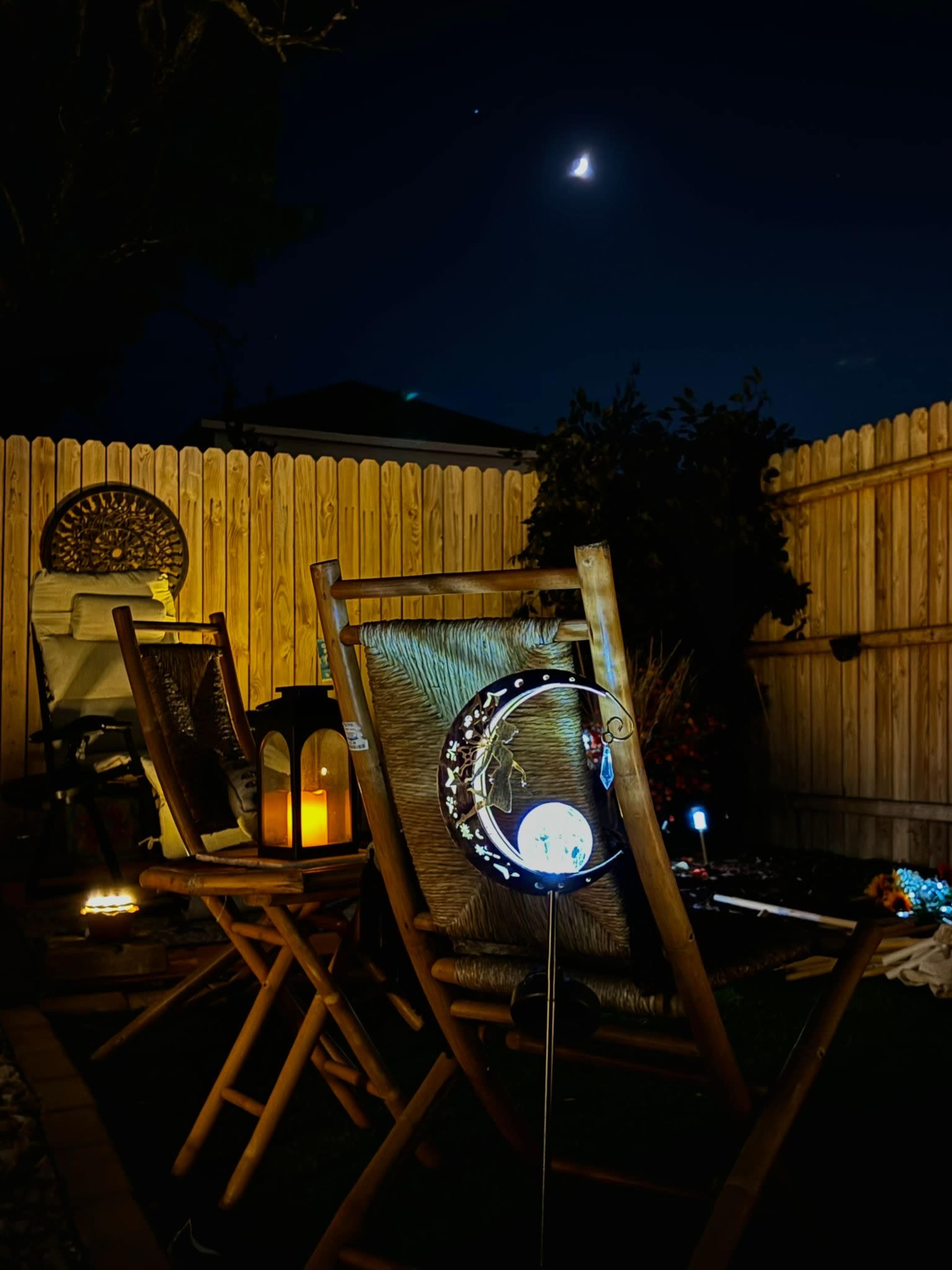 The image shows a backyard scene at night with two wooden chairs, a lantern on the ground, and a crescent moon visible in the sky.