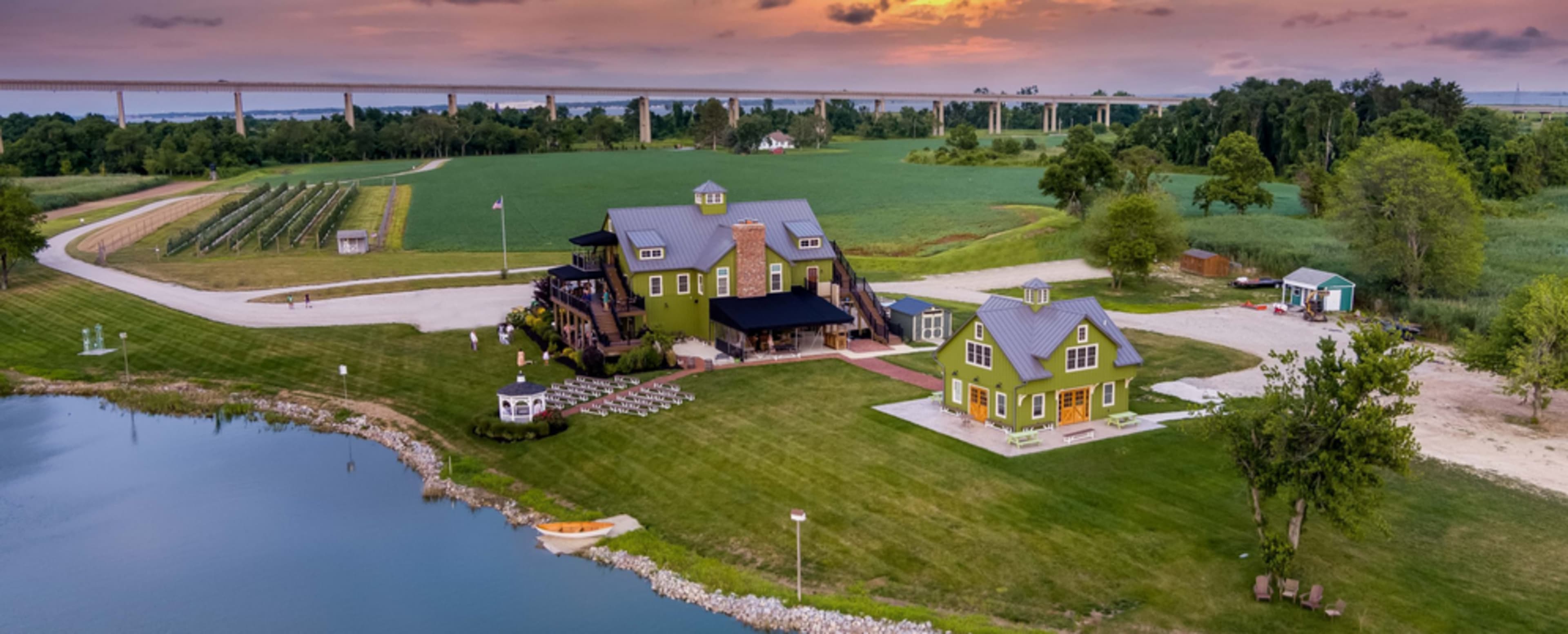 A green farmhouse with several outbuildings, set beside a pond and surrounded by fields under a cloudy sky.