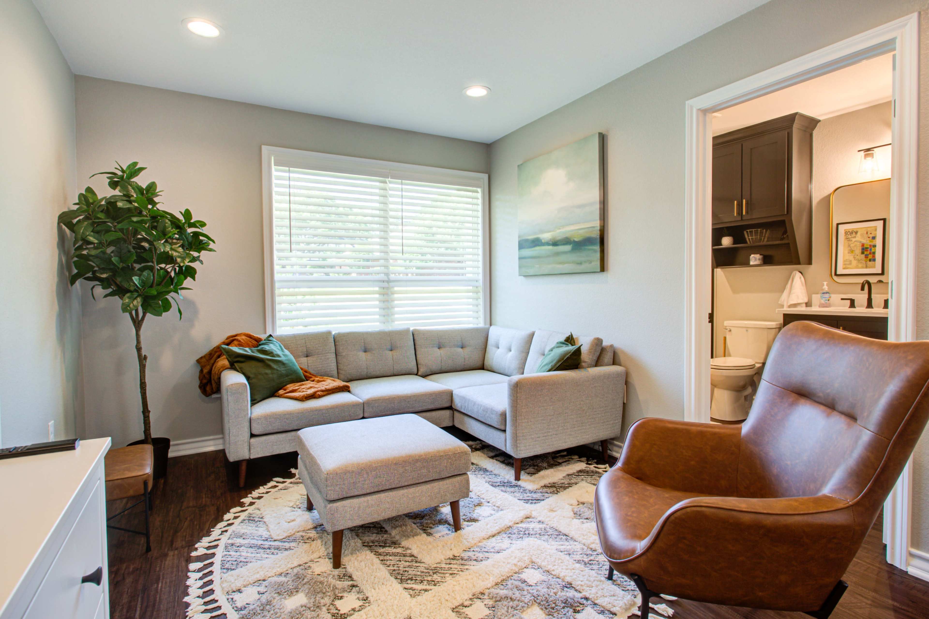 A modern living room features a grey sectional sofa, a brown leather armchair, a round patterned rug, and a large window with blinds.