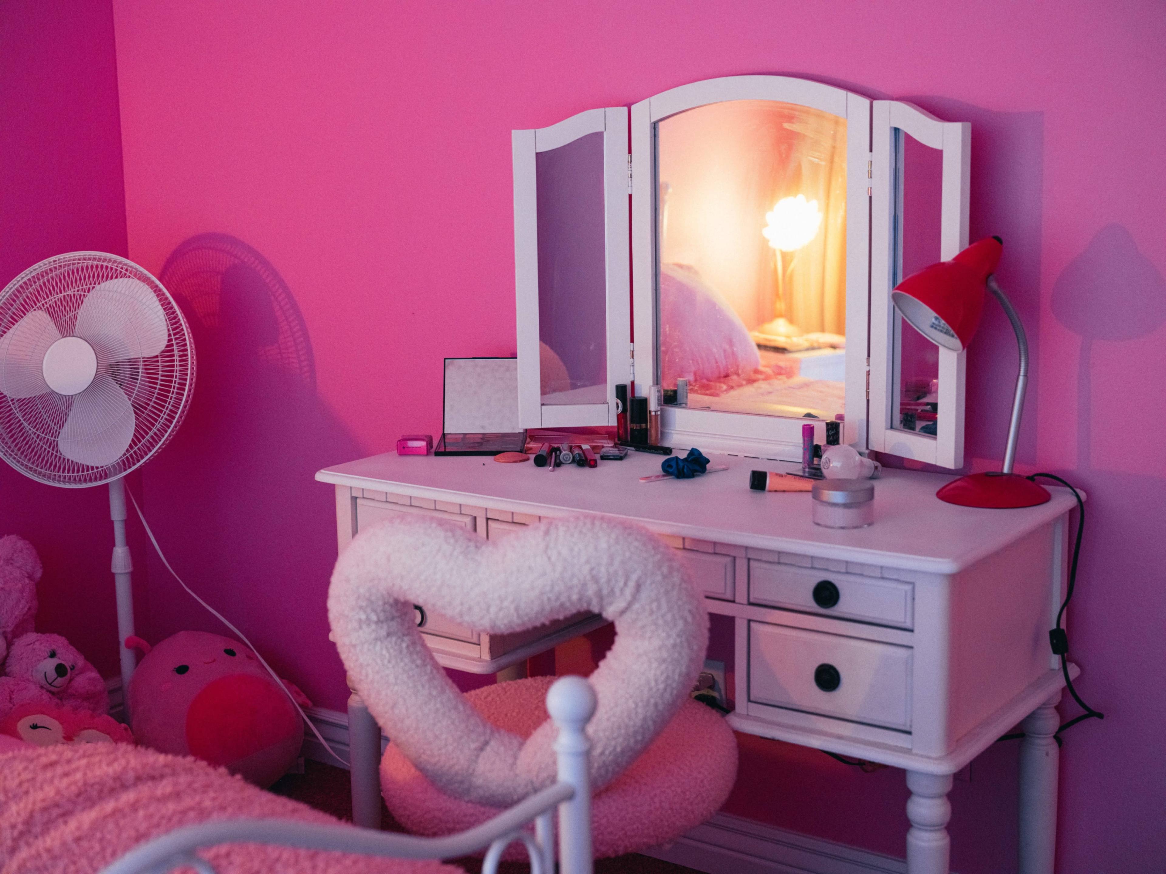 A white vanity sits against a pink wall, reflecting light from a lamp and surrounded by a fan and plush toys.