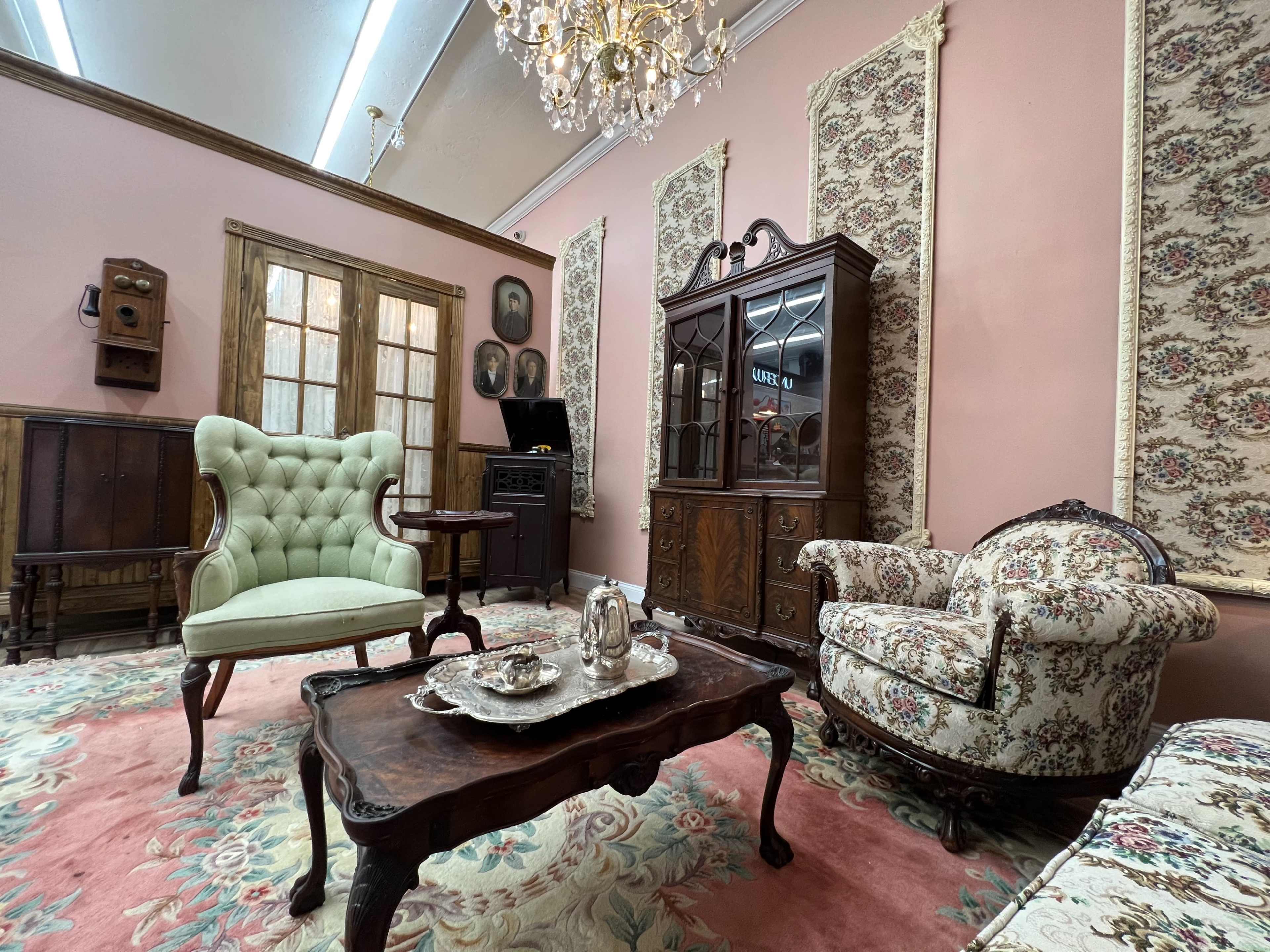 An elegantly furnished parlor with a green upholstered chair, ornate floral-patterned furniture, a wooden cabinet, and a vintage chandelier hanging from the ceiling.