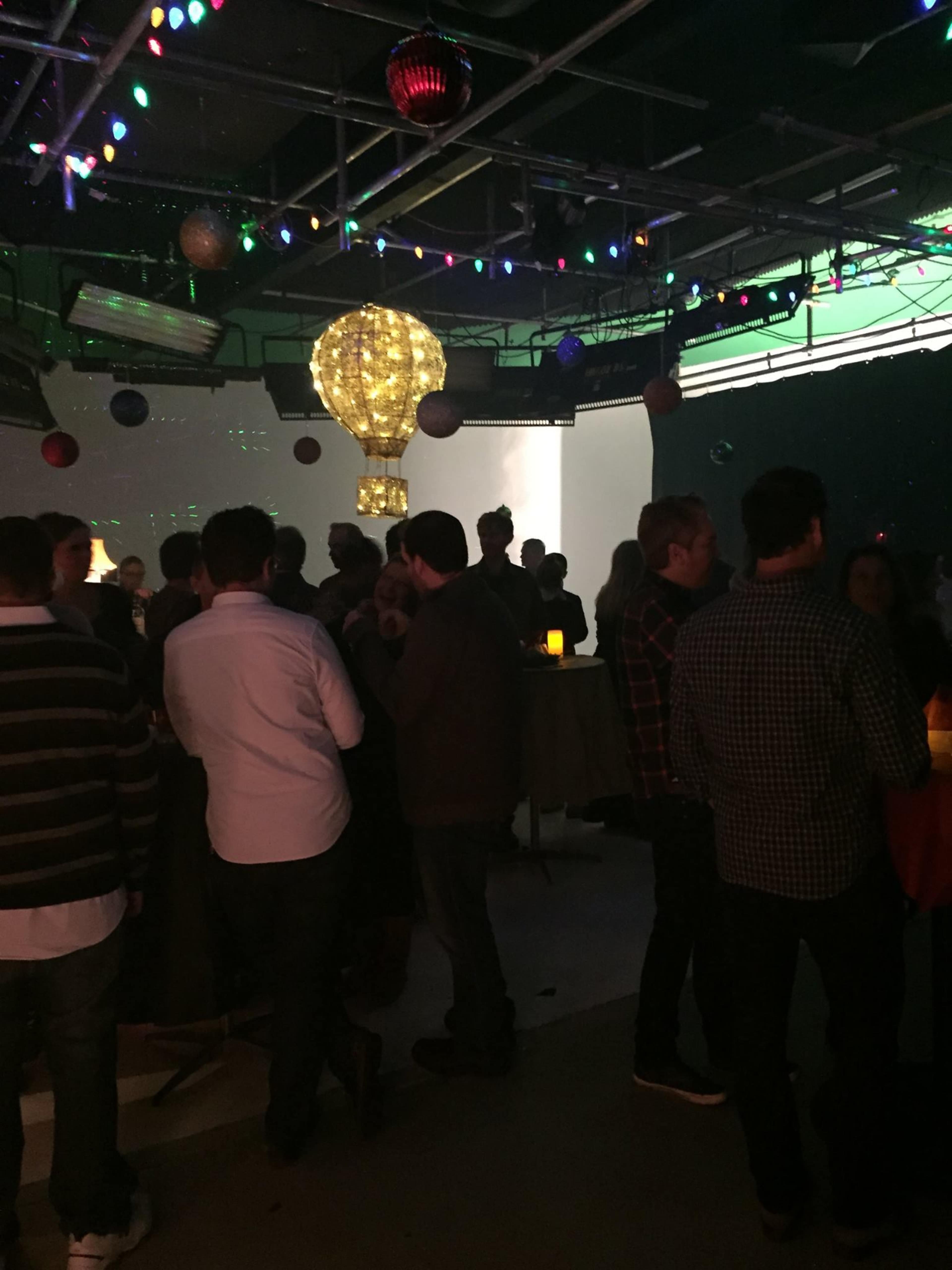 A lively gathering takes place indoors, with people mingling under colorful lights and a decorative hot air balloon hanging from the ceiling.