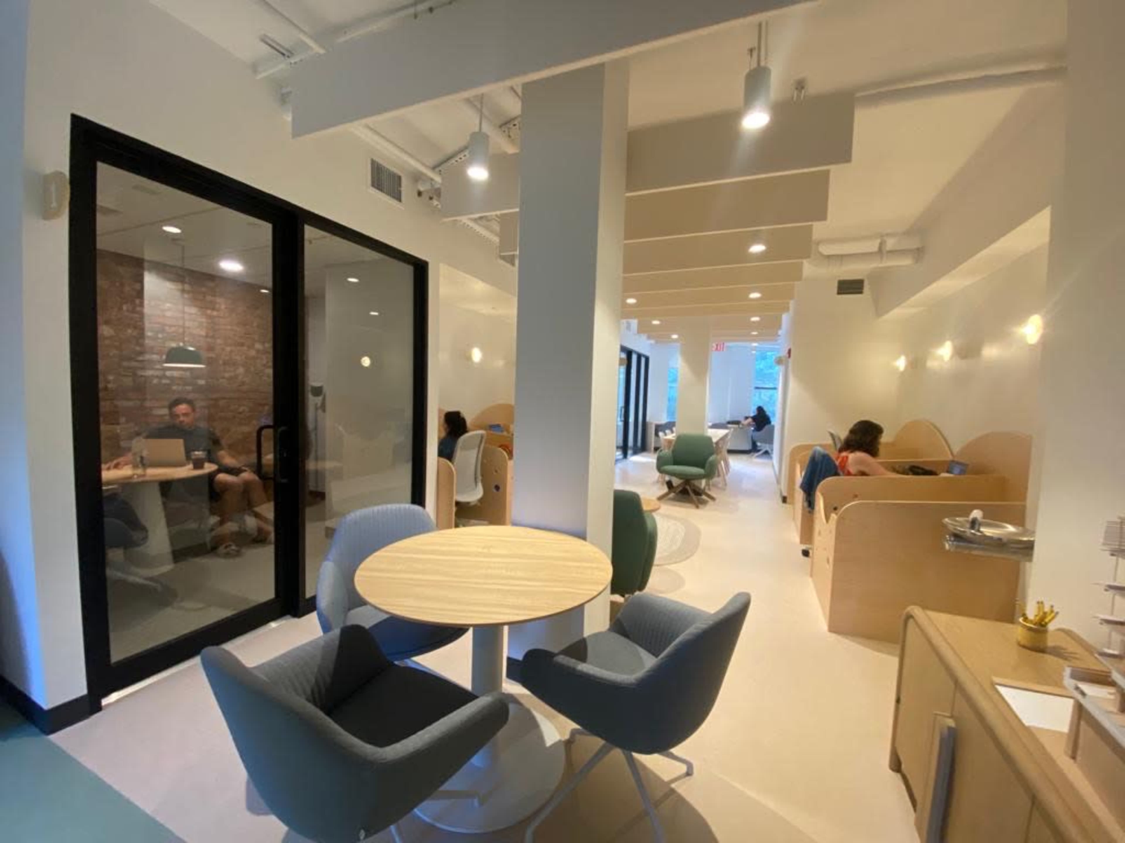 The image shows a modern coworking space with a mix of private meeting areas and open seating, featuring round tables and various seating arrangements.
