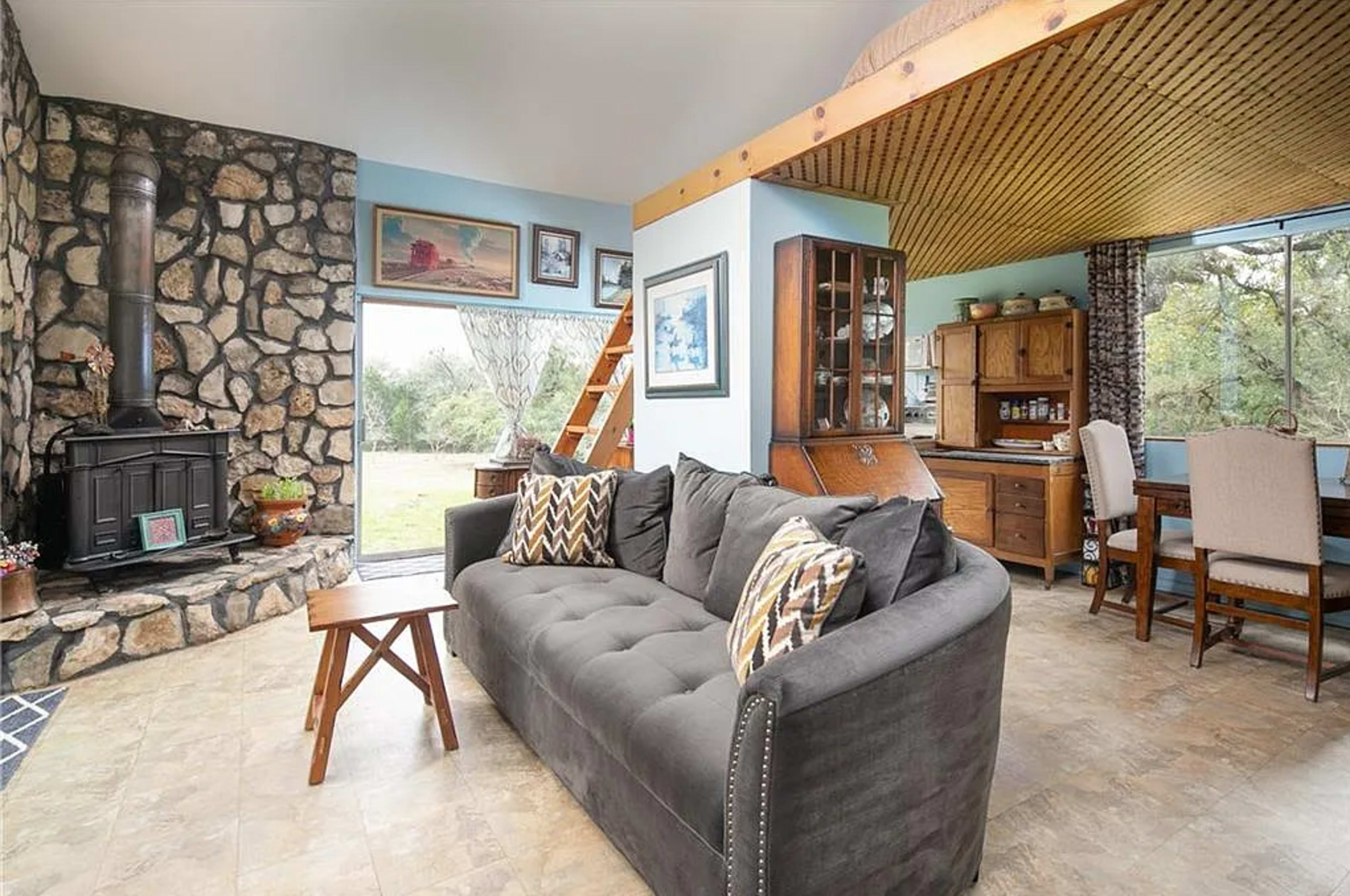 The image shows a cozy living space with a gray couch and a stone fireplace, adjacent to a dining area featuring wooden furniture and large windows that offer views of the outdoors.