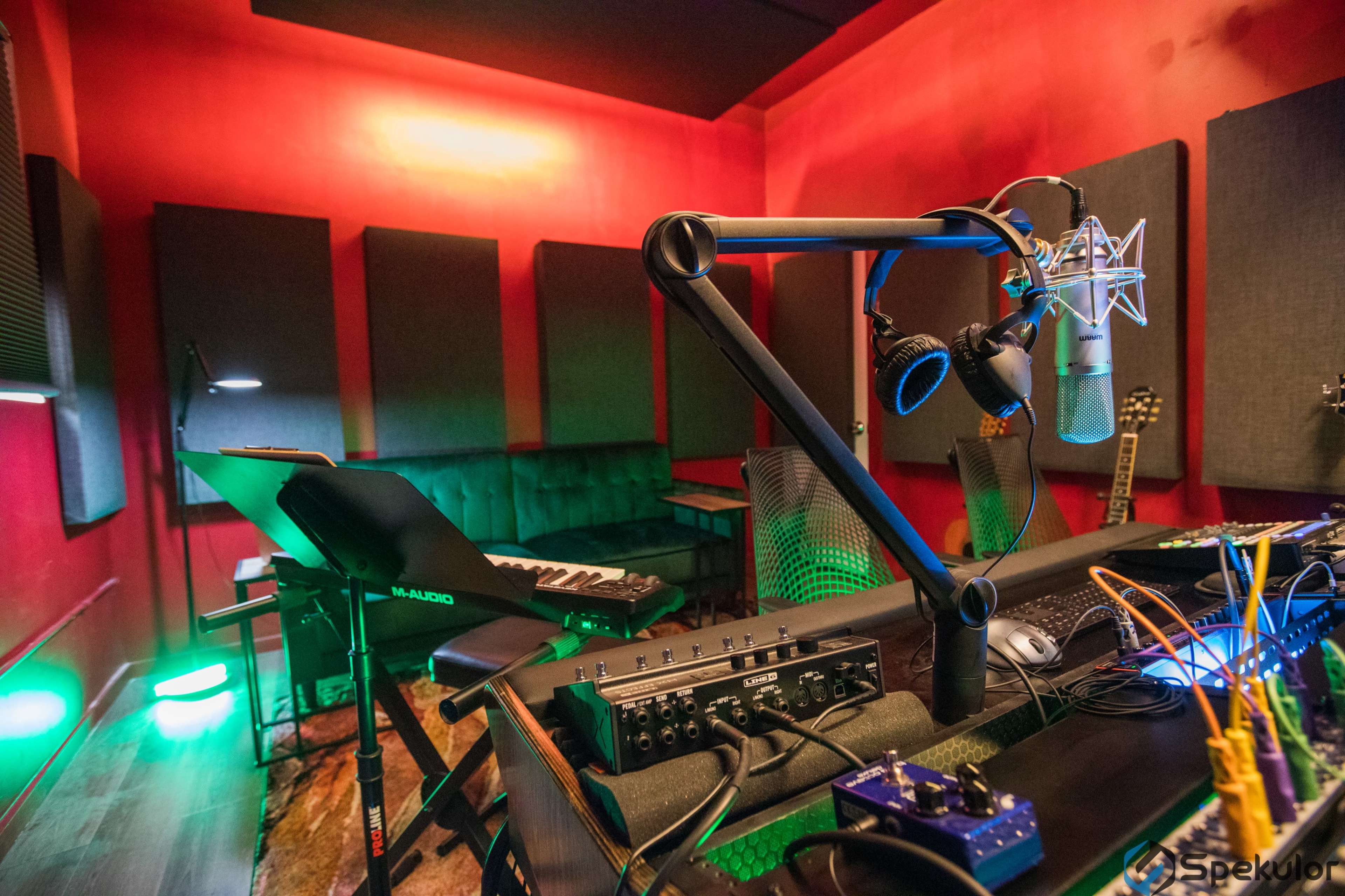A sound recording studio with red walls, equipped with a microphone, headphones, a keyboard, and various audio equipment arranged on a desk.