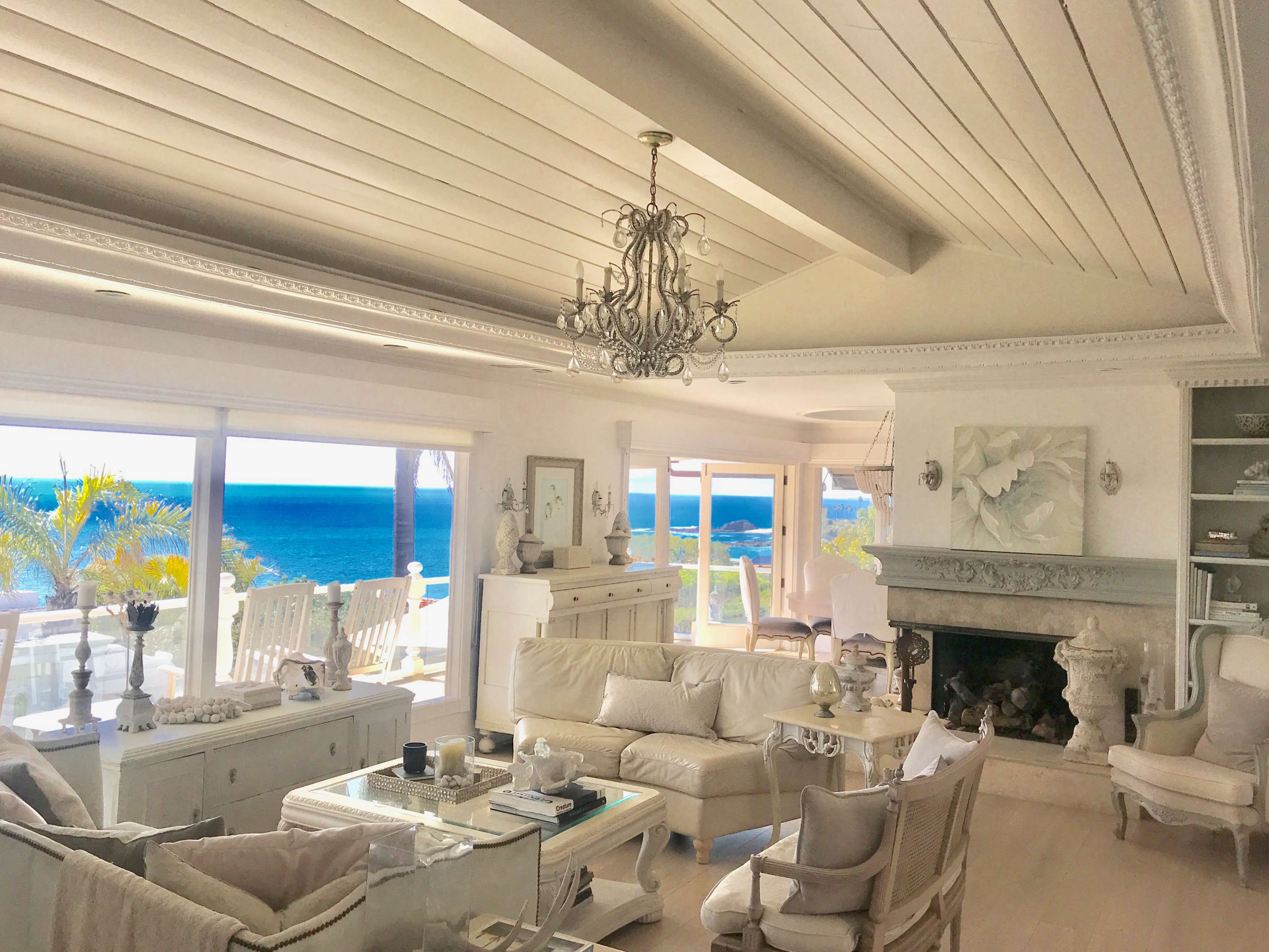 A bright living room features white furniture, a chandelier, and large windows overlooking the ocean.
