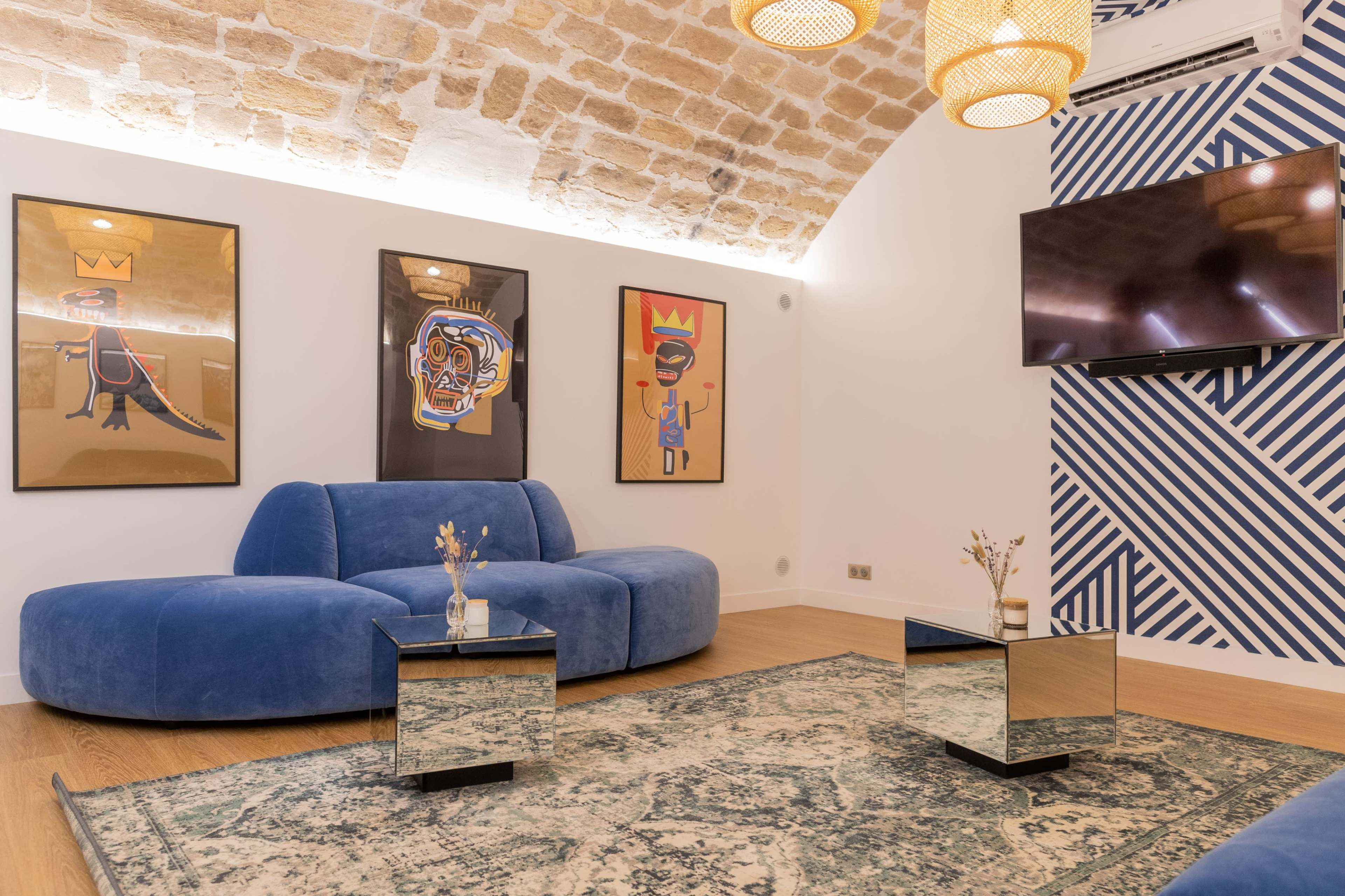 A modern living room featuring a blue curved sofa, two mirrored coffee tables, and colorful framed artwork on the walls, with a television mounted in the corner.