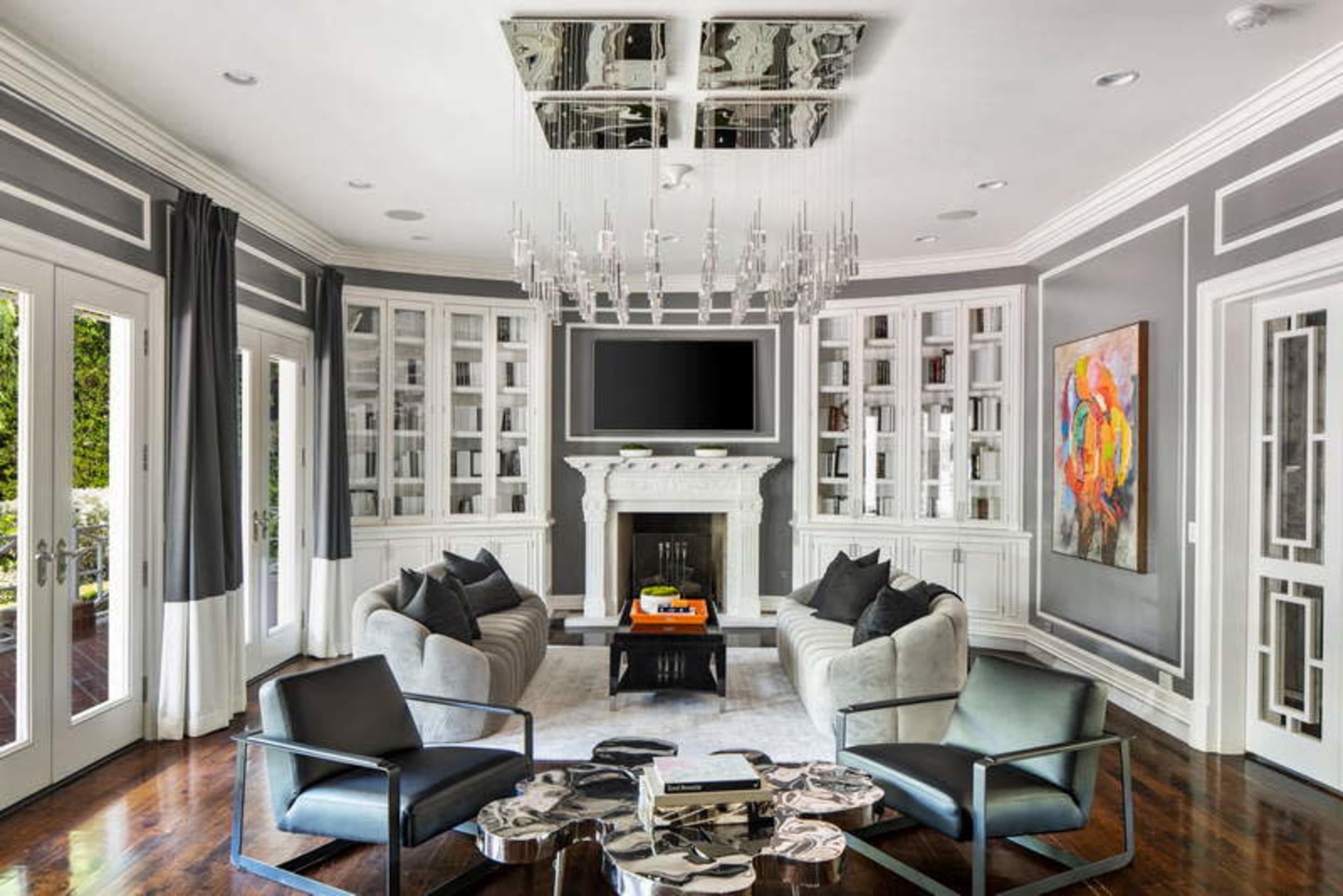 A modern living room with a fireplace, large windows, a chandelier, and sleek furniture arranged around a central coffee table.