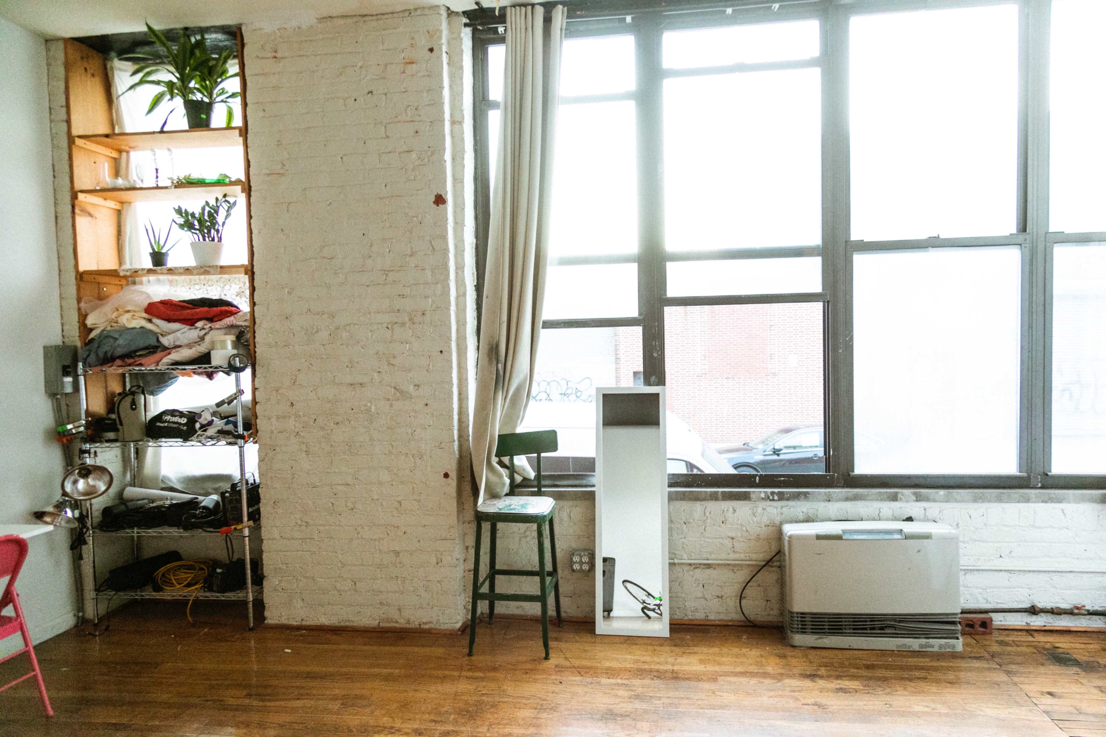 The image shows a sparsely furnished room with large windows, a green chair, a white column, and shelves displaying plants and various items.