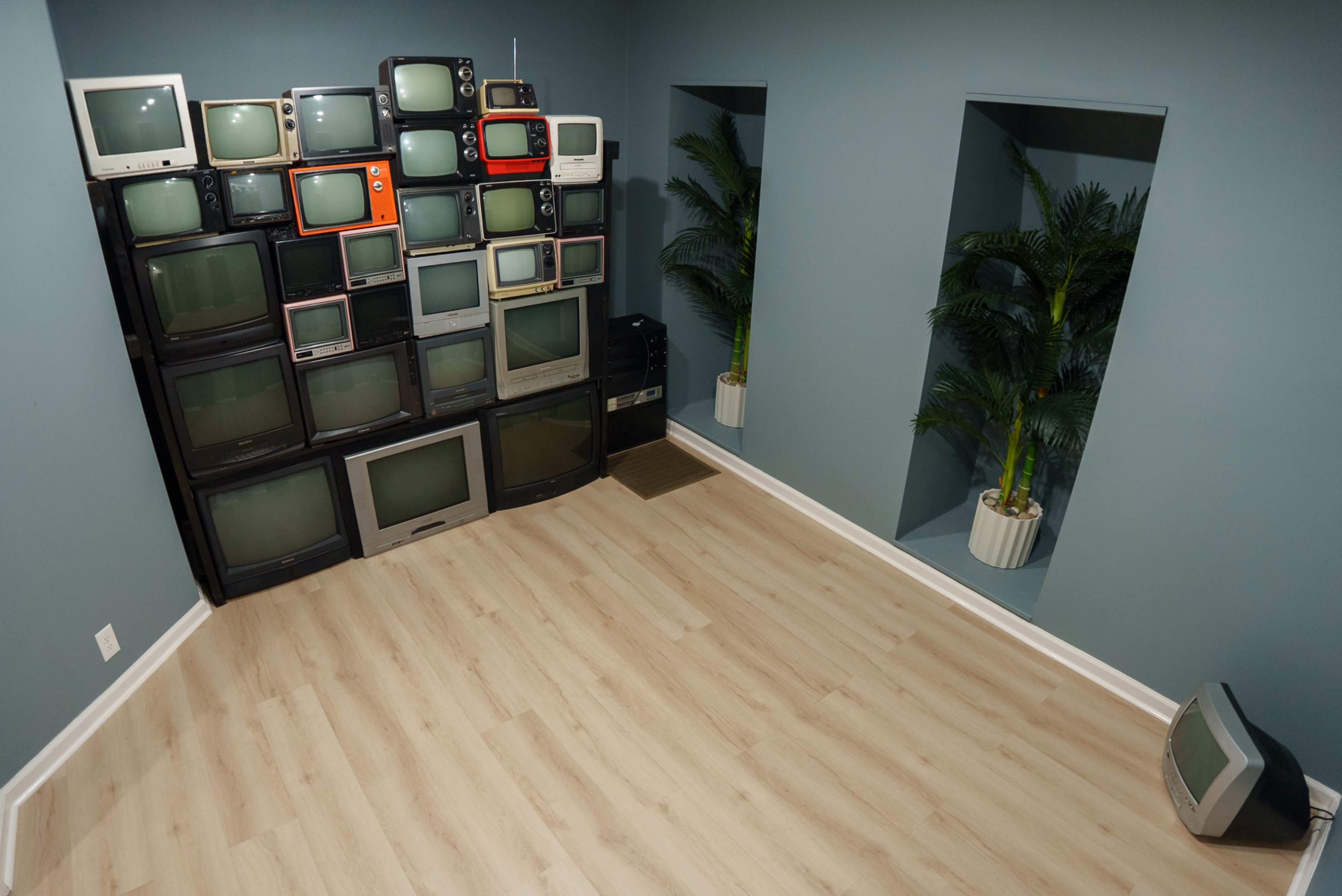 The image shows a room with a wall of stacked vintage televisions and two potted plants in alcoves.