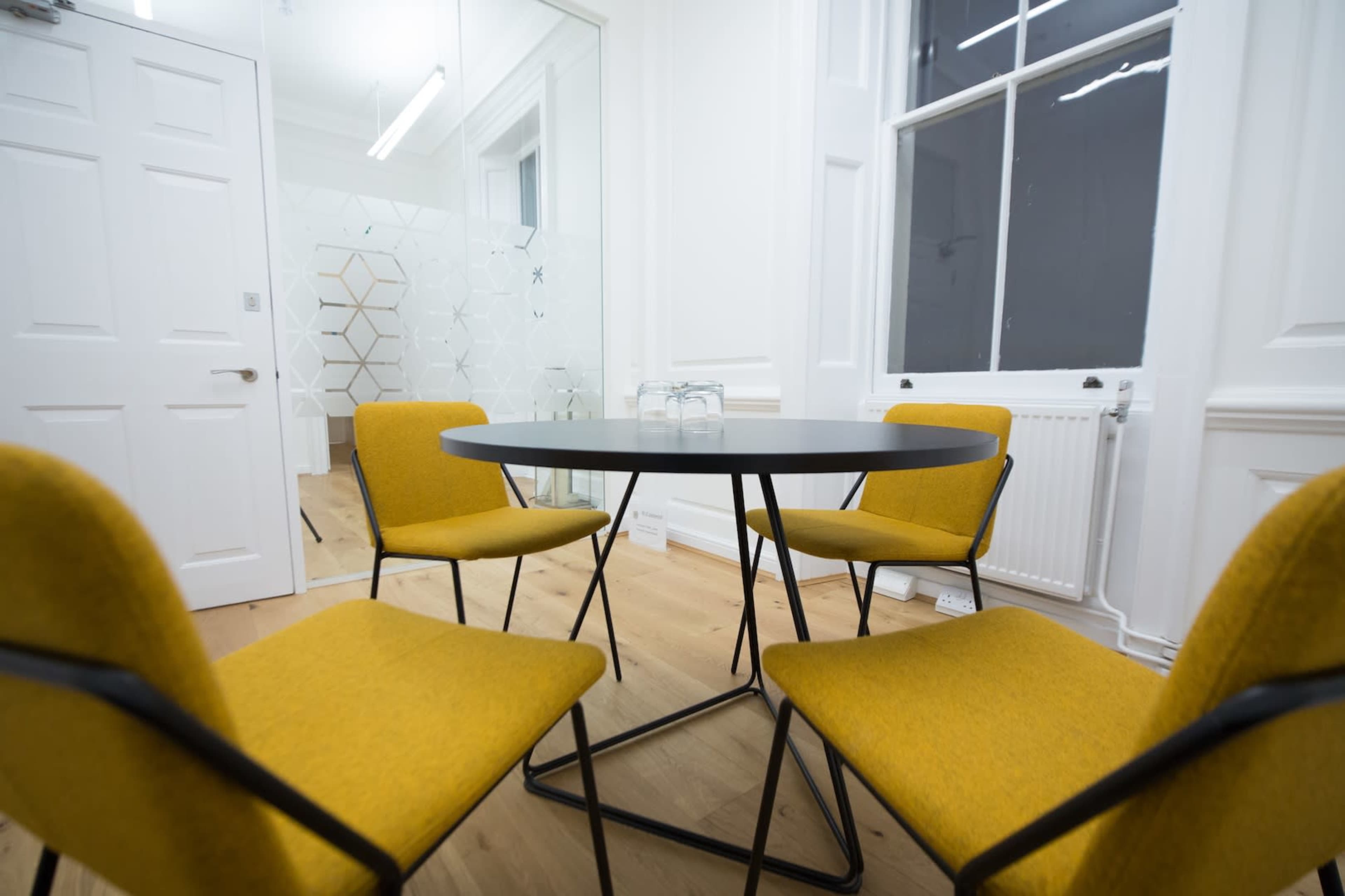 A round table is surrounded by four yellow chairs in a well-lit room with white walls and a large window.