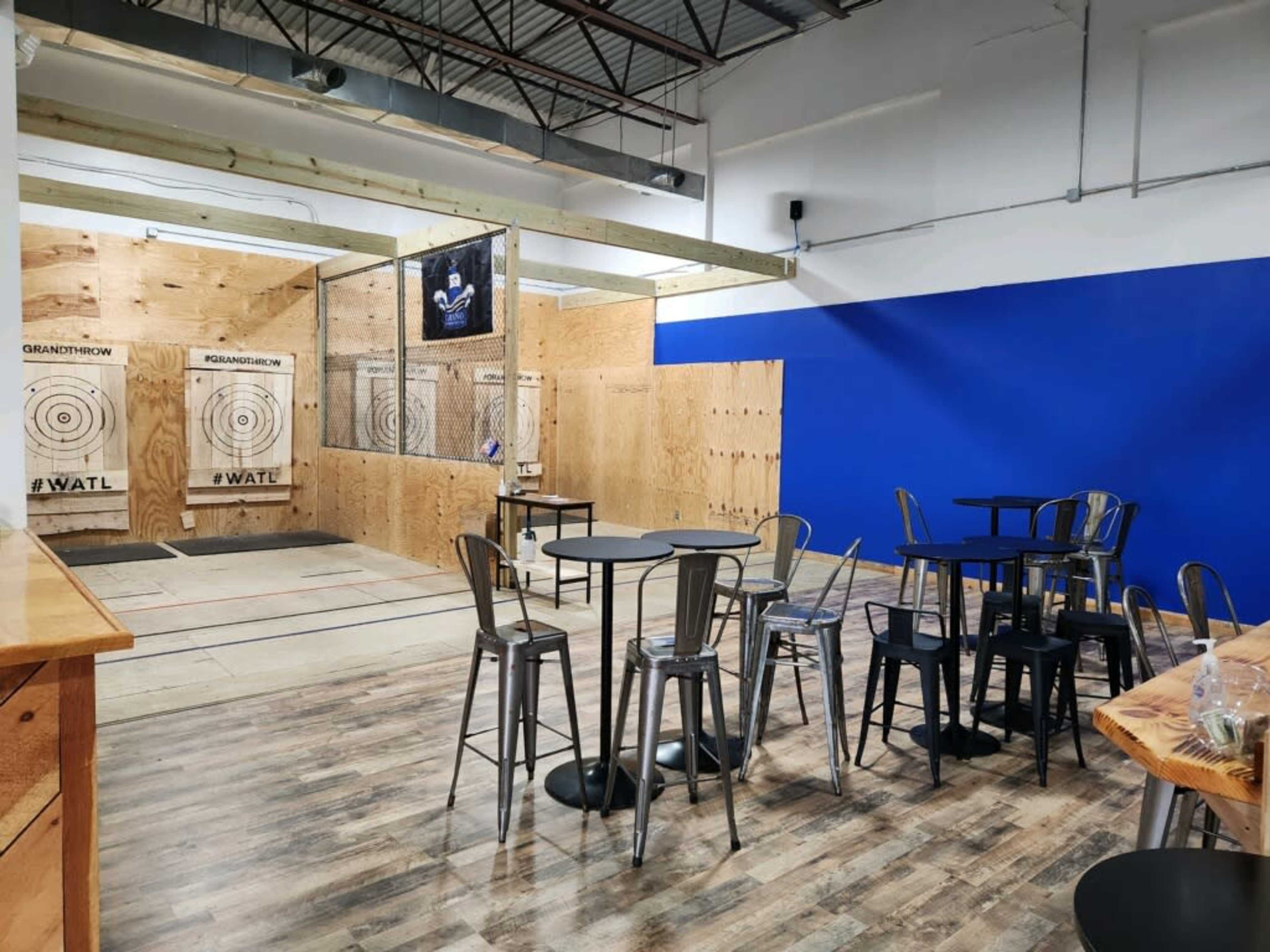 A spacious indoor venue featuring a wooden floor, multiple axe-throwing targets, and seating areas with metal tables and chairs.