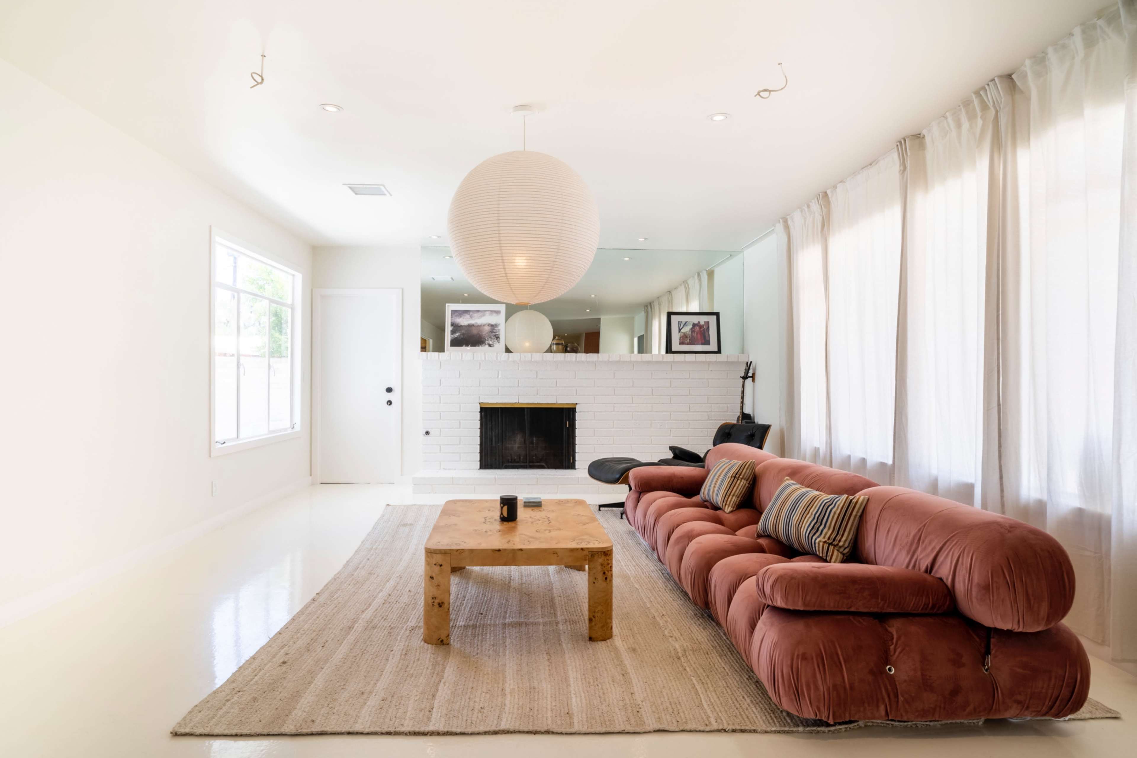 A spacious living room features a pink sectional sofa, a wooden coffee table, and a white brick fireplace surrounded by large windows and soft curtains.