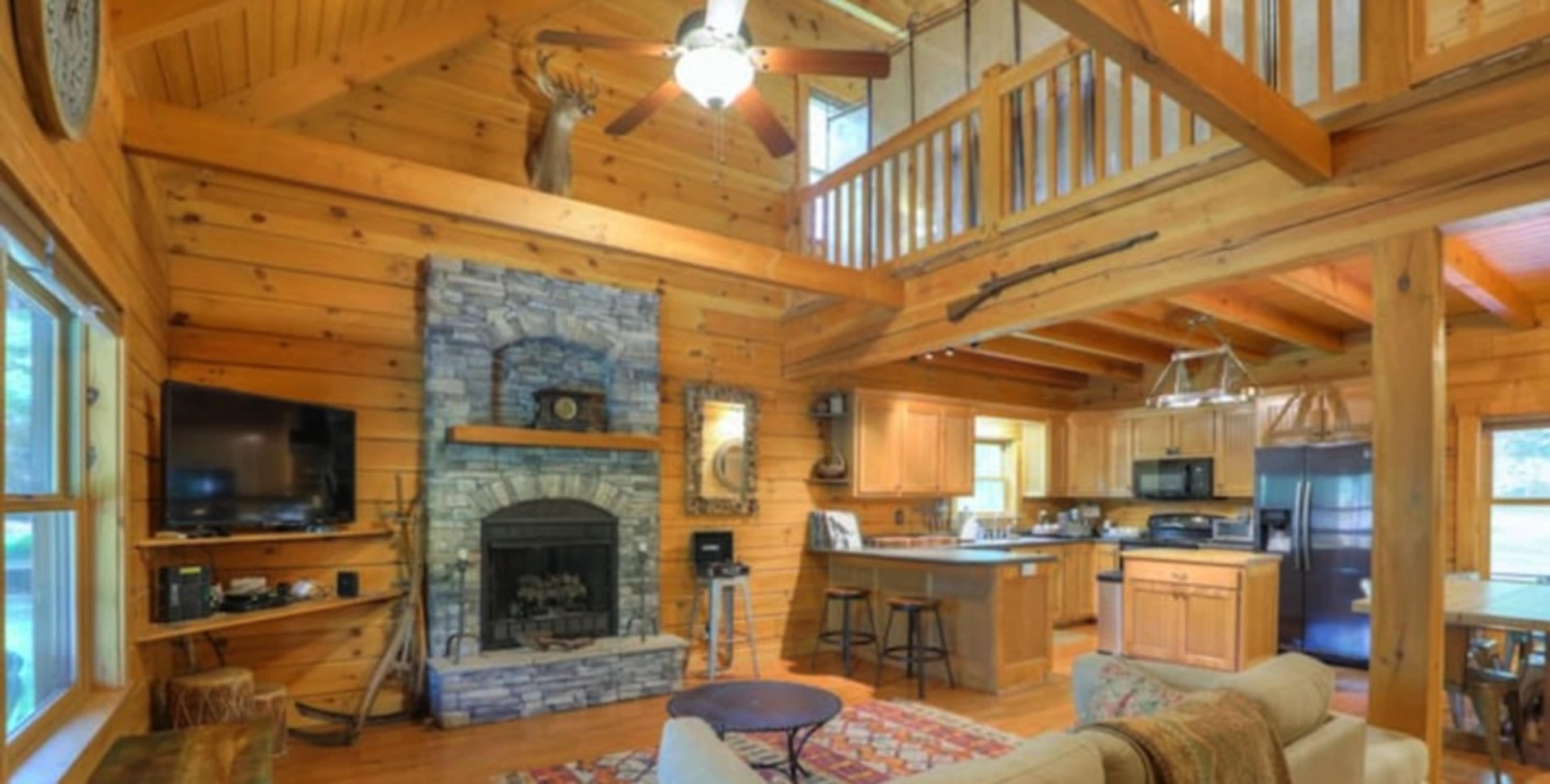 A rustic wooden cabin interior with a stone fireplace, a spacious living area, and an open kitchen visible in the background.