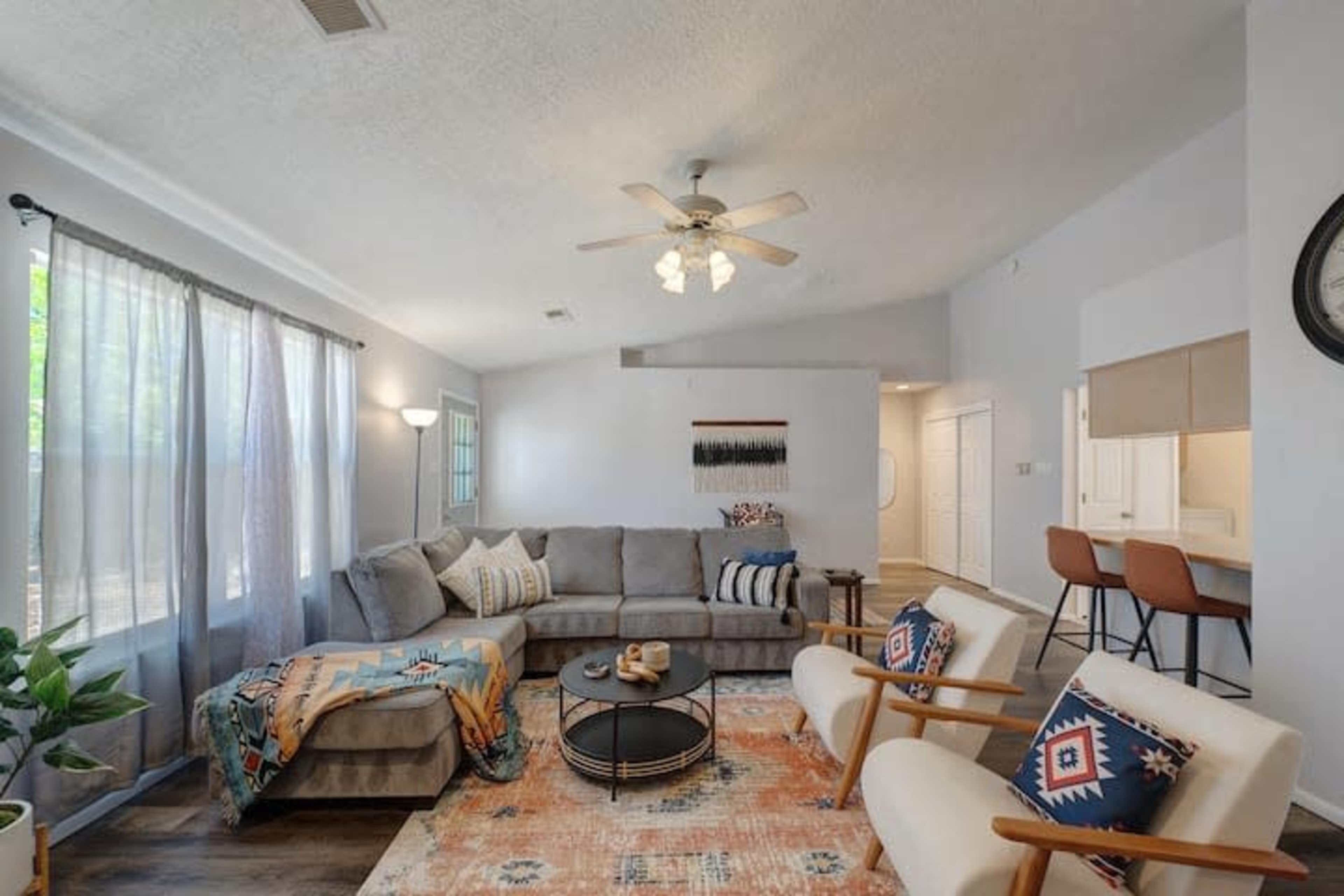 A cozy living room featuring a gray sectional sofa, a round coffee table, two armchairs, and a large window with sheer curtains.