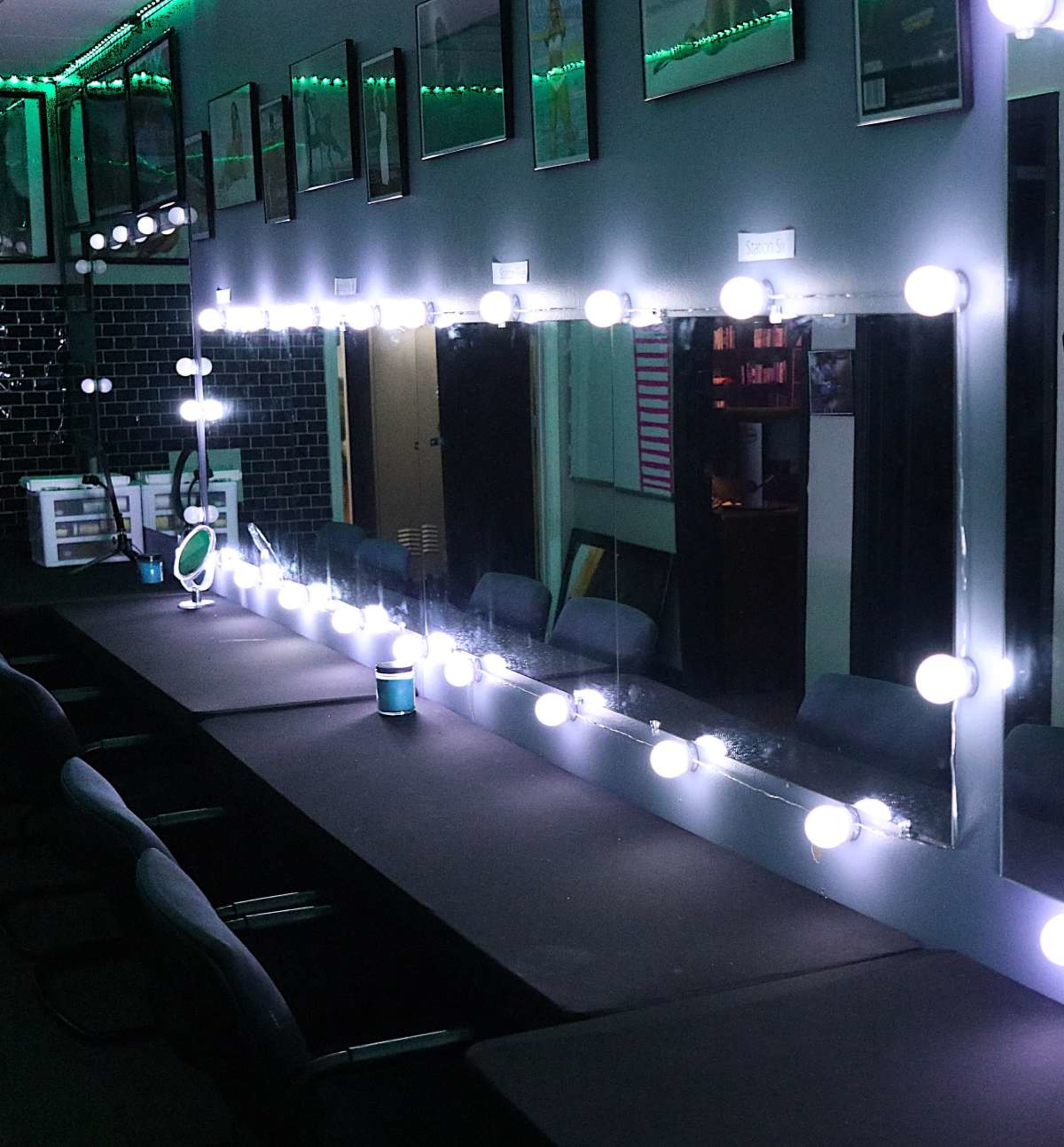 The image shows a well-lit makeup vanity with a large mirror surrounded by illuminated bulbs, set against a dark wall with framed pictures.