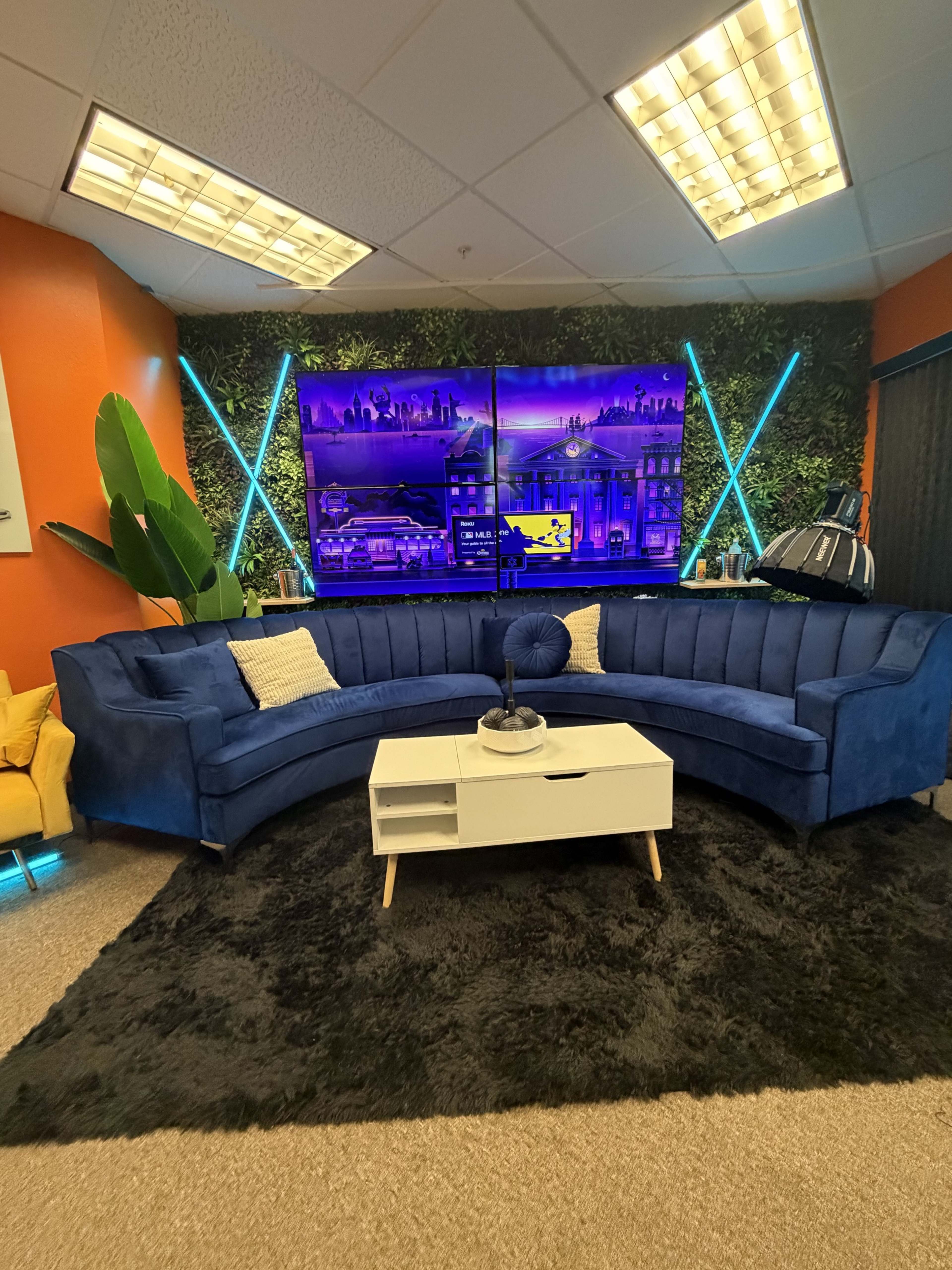 The image shows a modern lounge area with a blue curved sofa, a white coffee table, a black rug, and a vibrant wall-mounted display featuring cityscapes.