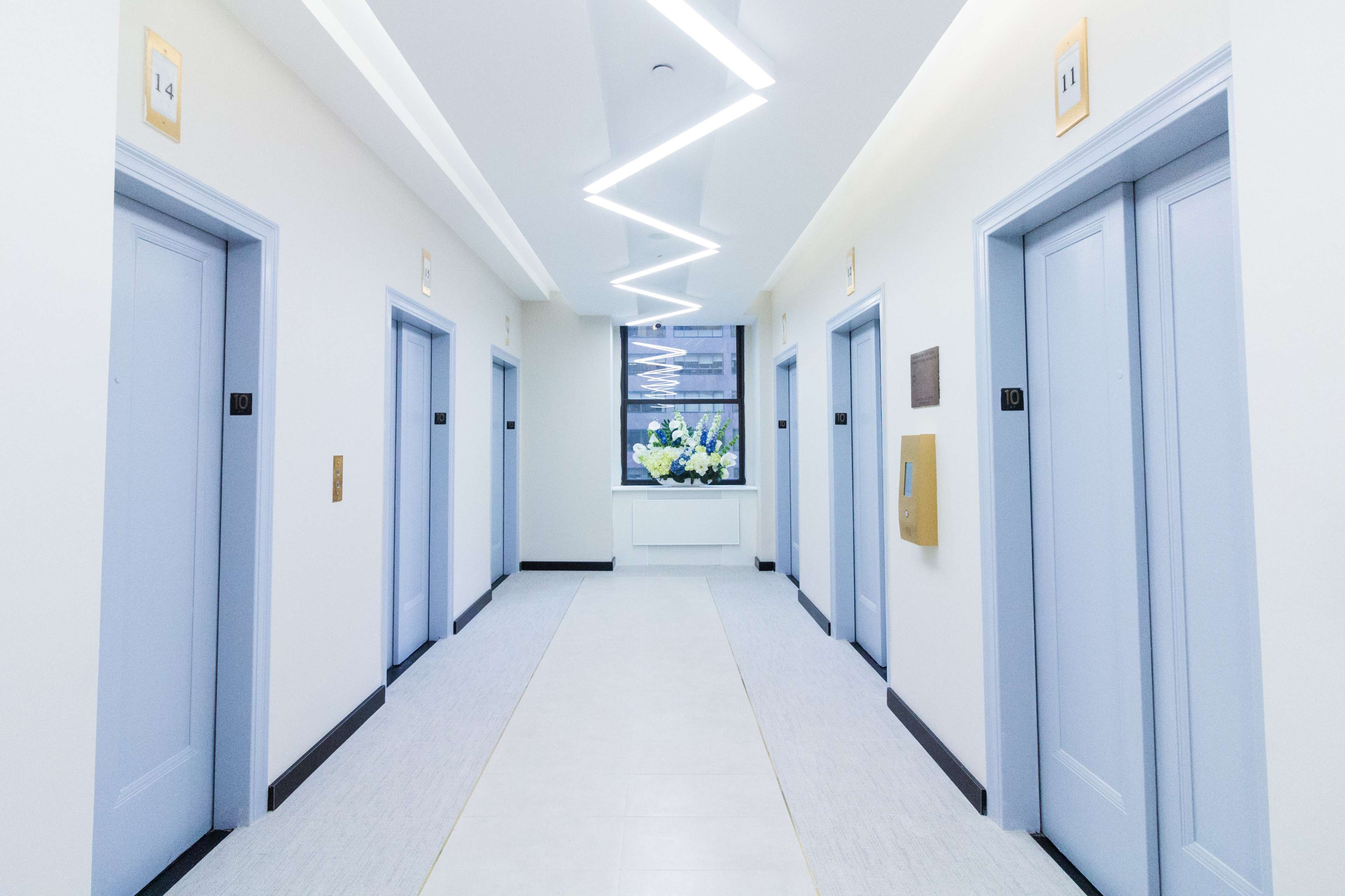 A long hallway features six elevator doors on either side, with a window at the end providing natural light.