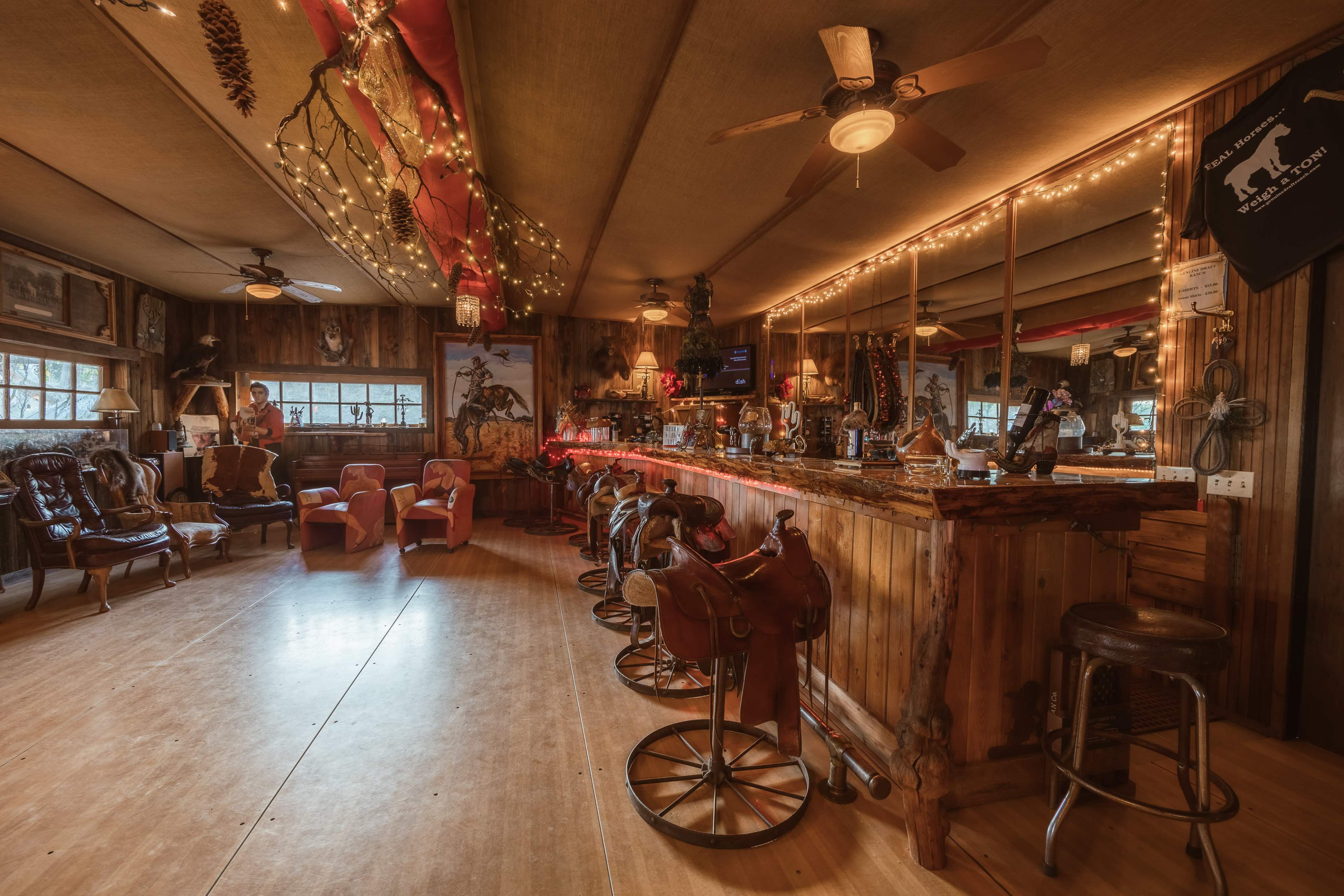 The image shows a rustic bar area in a lodge, featuring wooden walls, leather chairs, and string lights, with a counter made of polished wood and saddle-shaped stools.