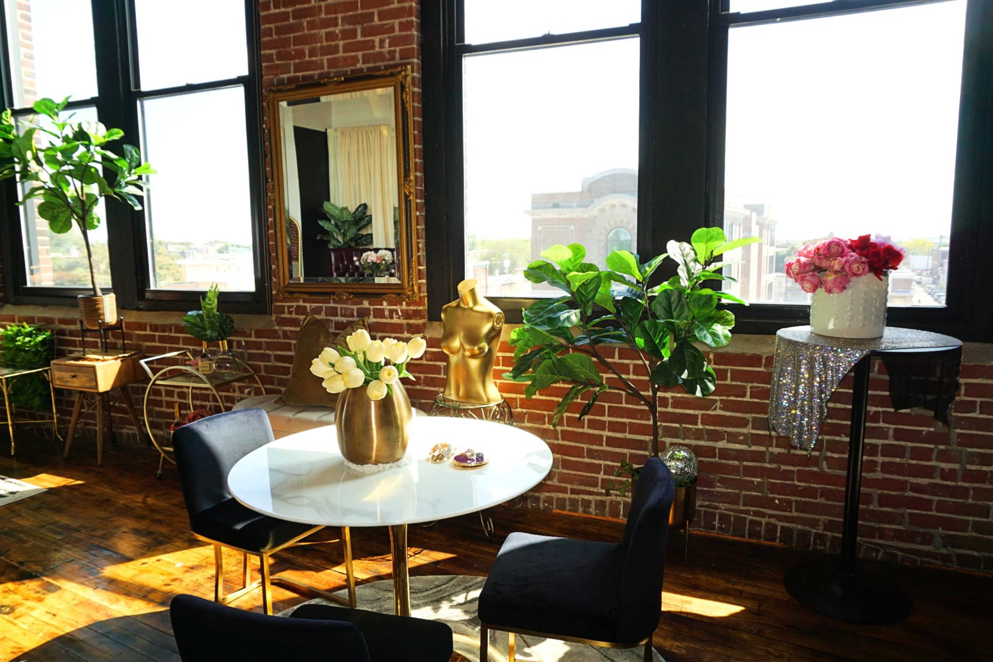 A modern interior space with a round white table, black chairs, houseplants, and a decorative mannequin against a backdrop of large windows and exposed brick walls.