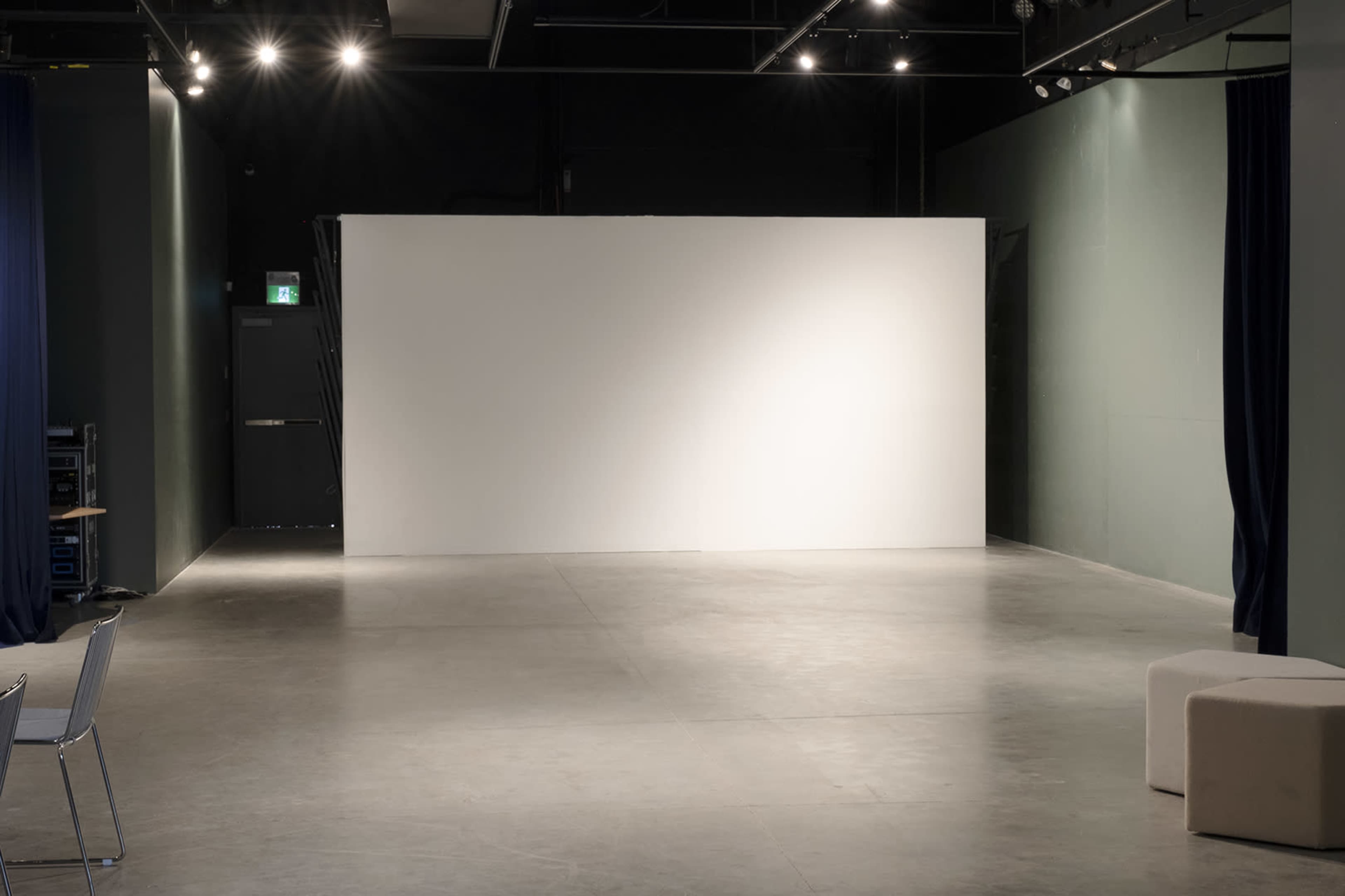 The image shows an empty performance space with a bare white wall and minimal furnishings.