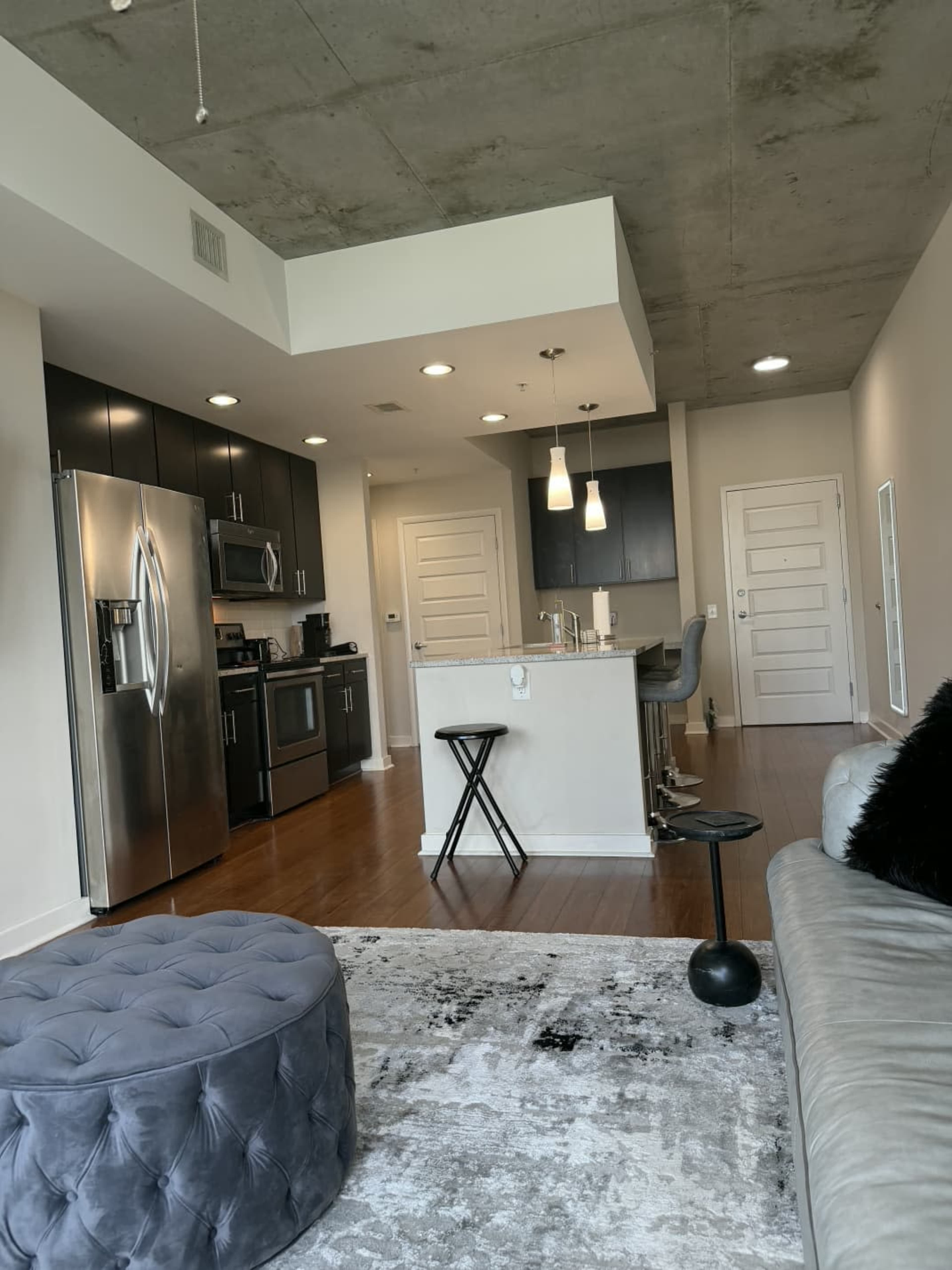 The image depicts a modern studio apartment featuring a kitchen with dark cabinetry, stainless steel appliances, a small dining area, and a seating area with a sofa and ottoman.
