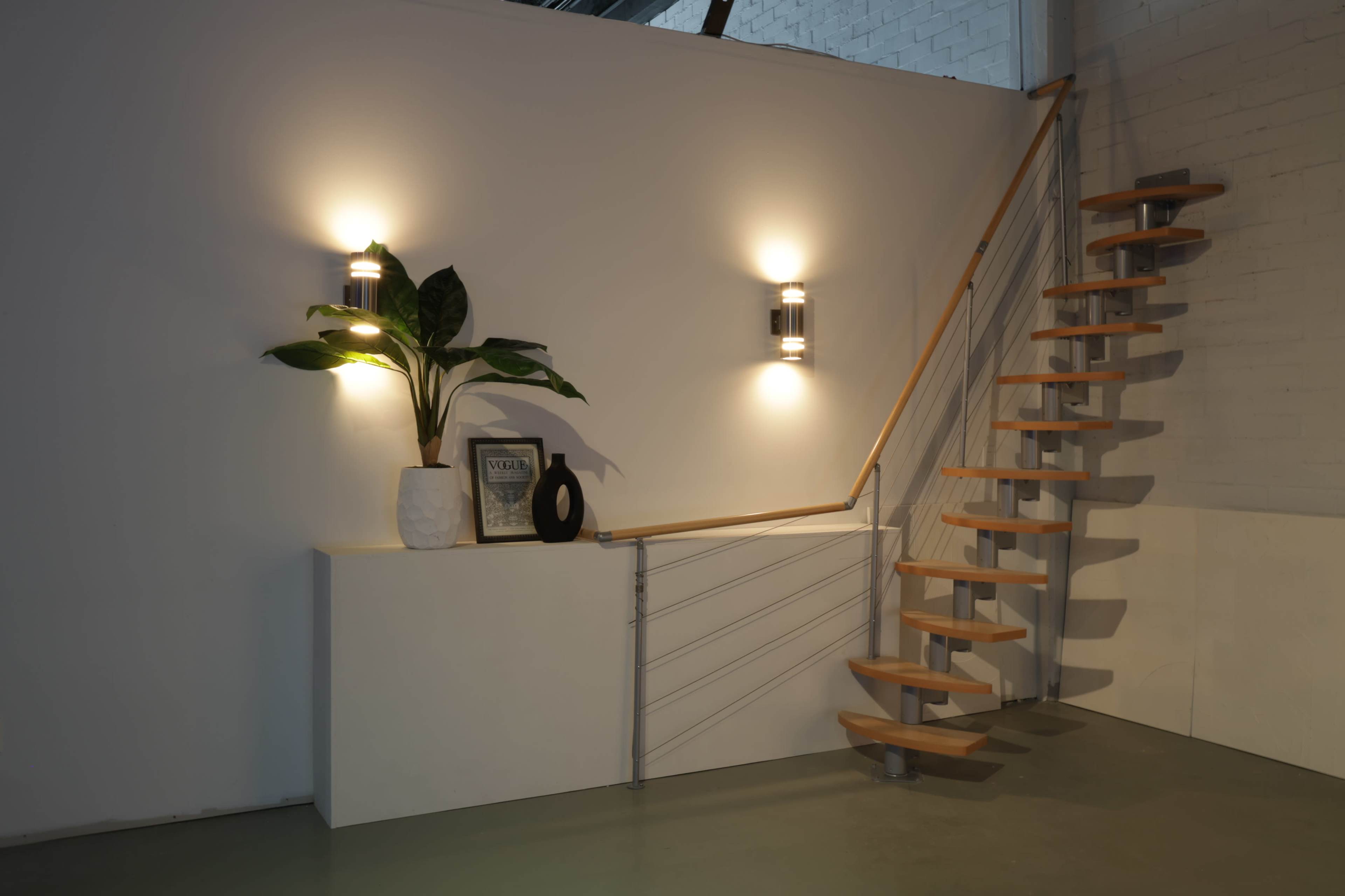 A modern interior features a wooden spiral staircase alongside a minimalist wall with two sconces, a plant, and decorative items on a sleek console.