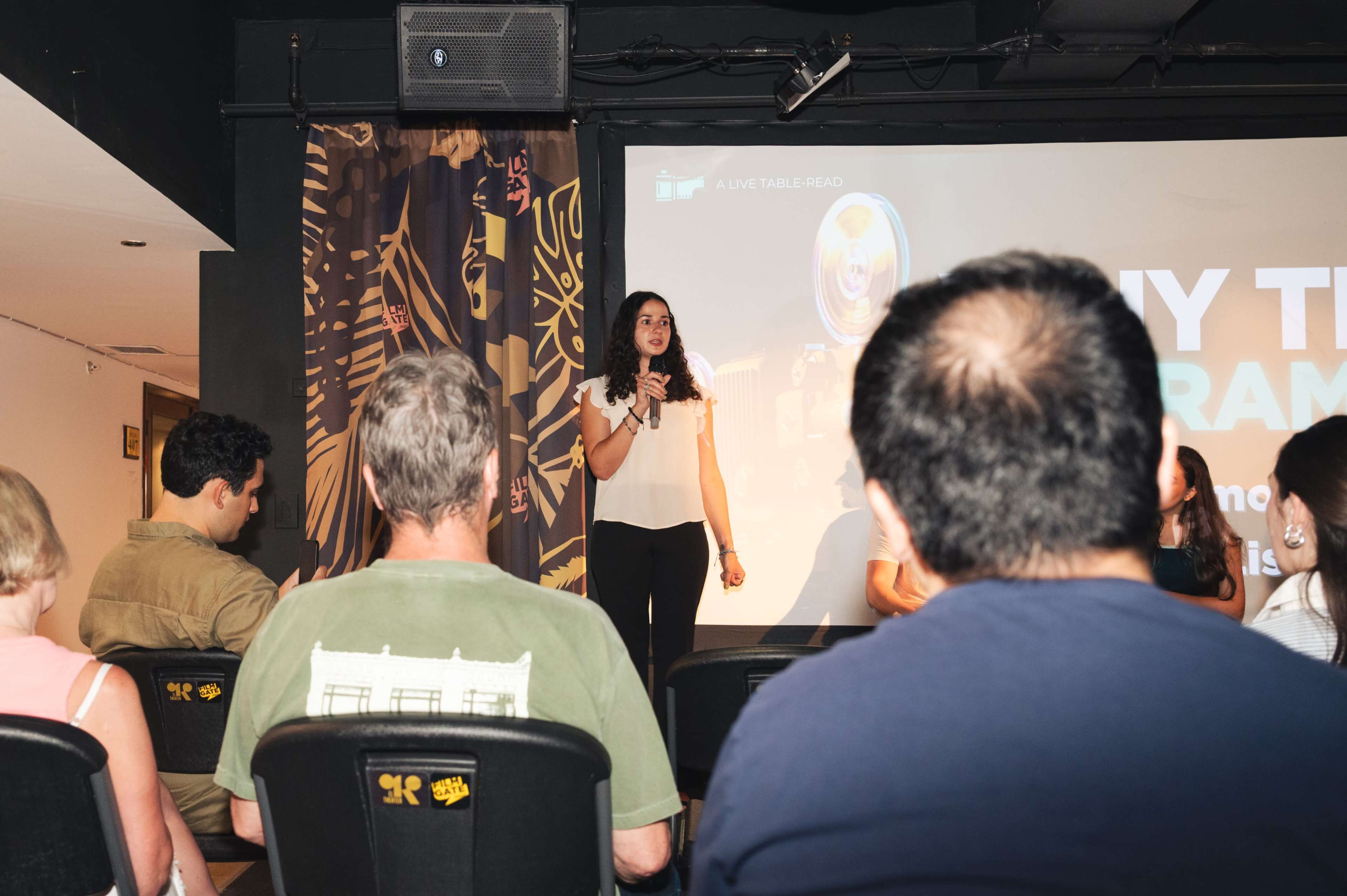 A woman stands on stage in front of an audience, presenting with a screen displaying the text "Why the Drama?" in the background.