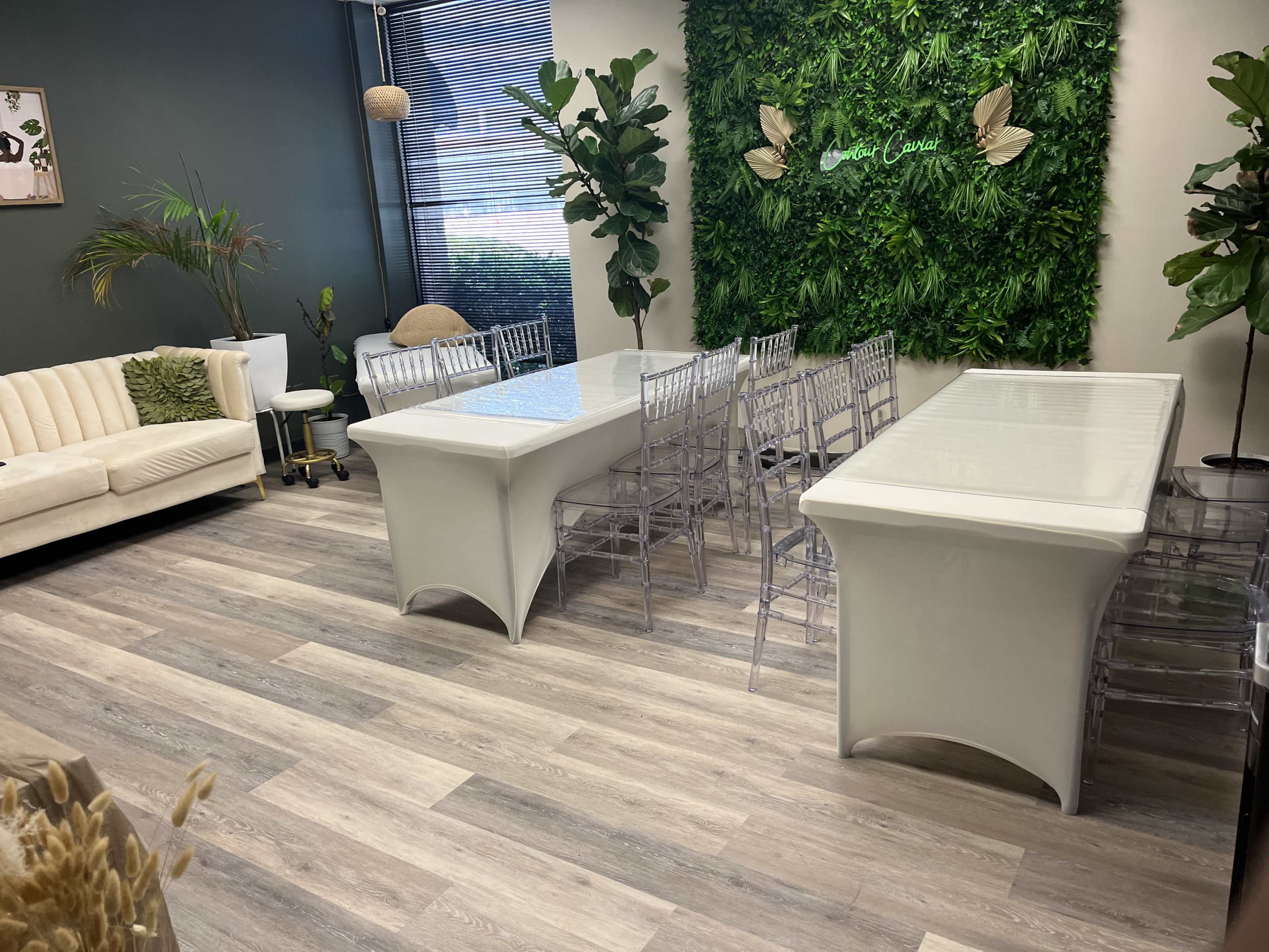 A modern room with two white tables, clear chairs, a sofa, and greenery on the walls.