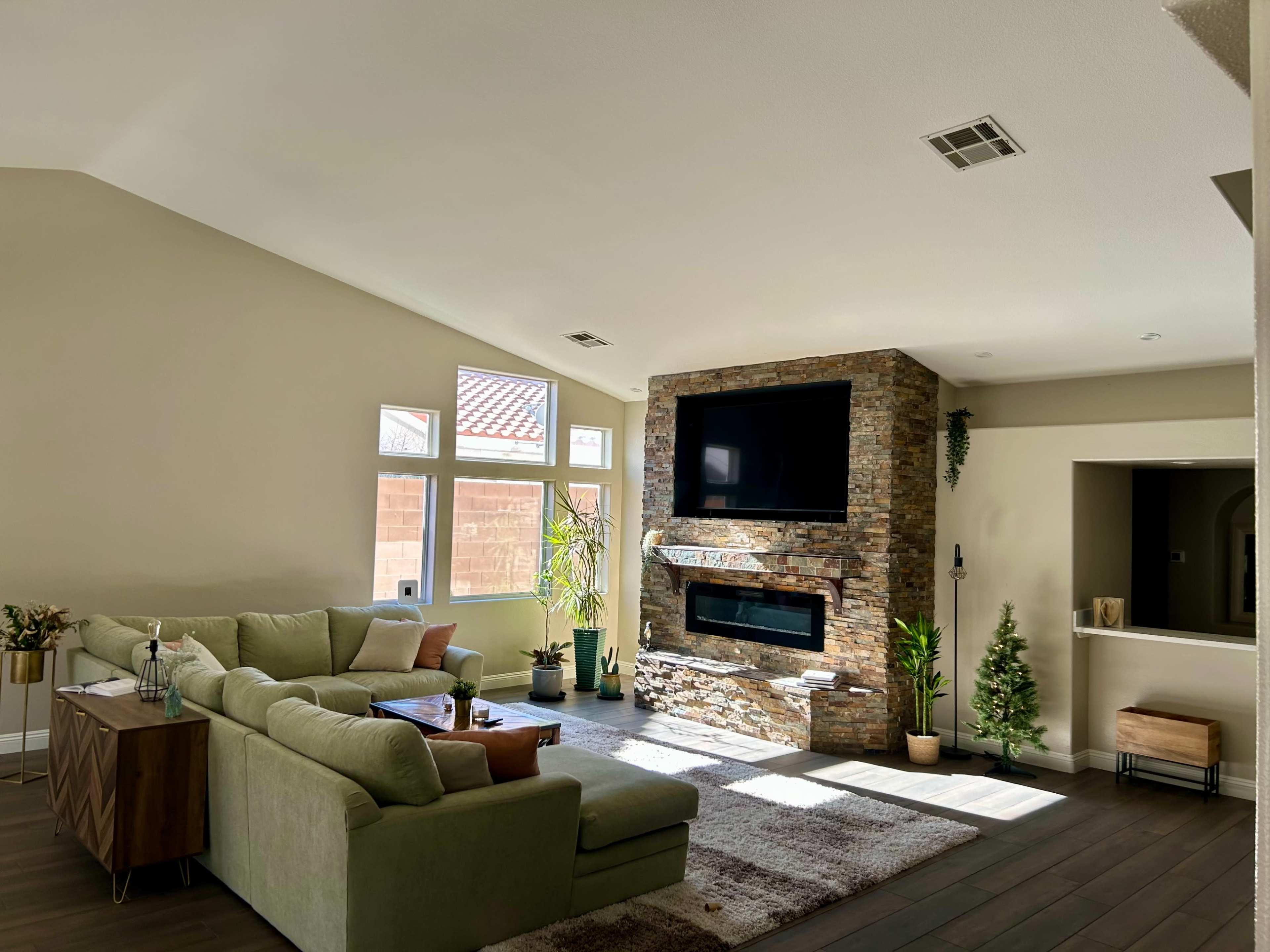 The image shows a spacious living room with a stone fireplace, a large television mounted on the wall, a sectional sofa, and several plants near the windows.