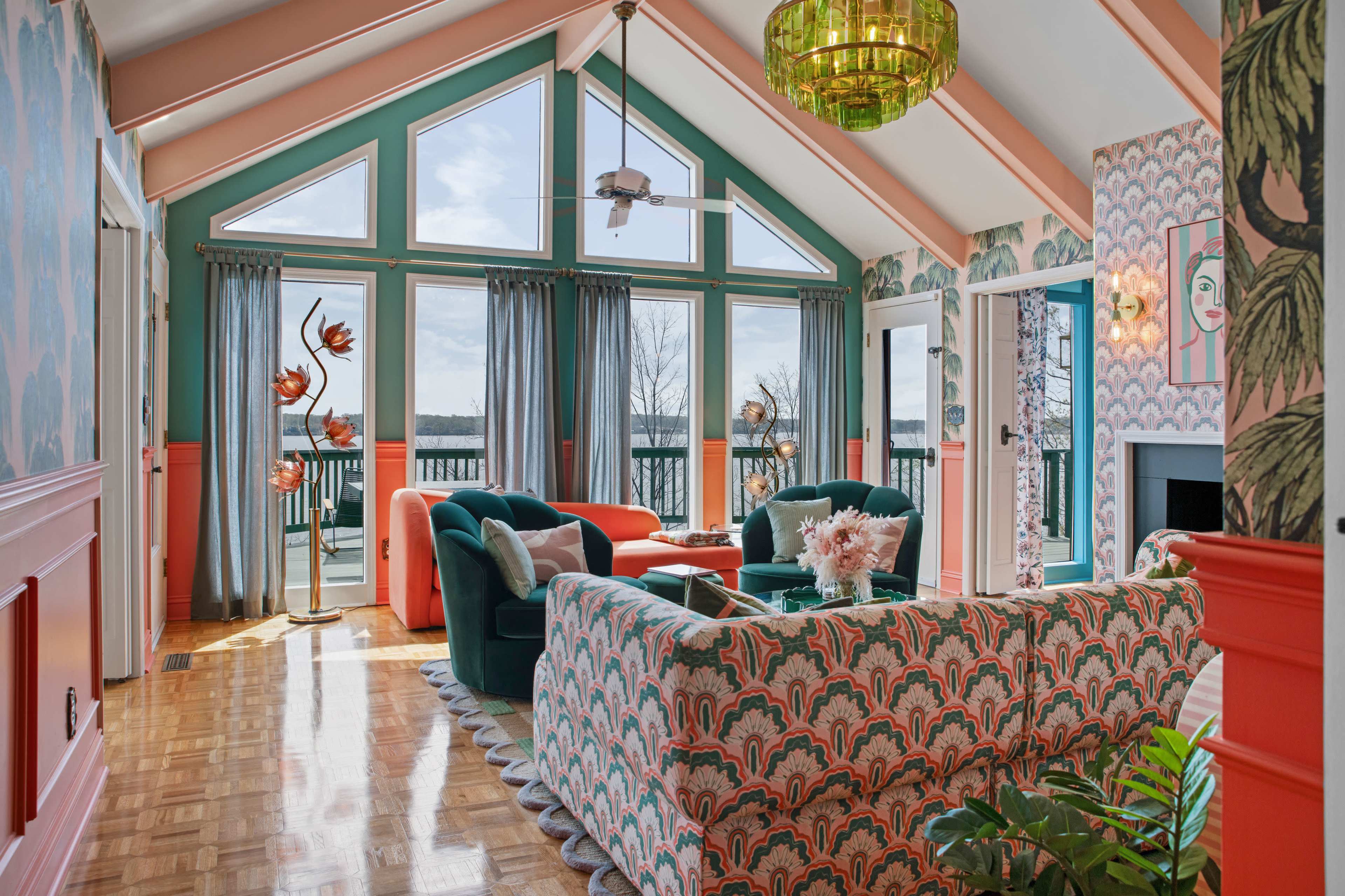 A brightly decorated living room features large windows with a view and colorful furniture, including a patterned sofa and green armchairs.