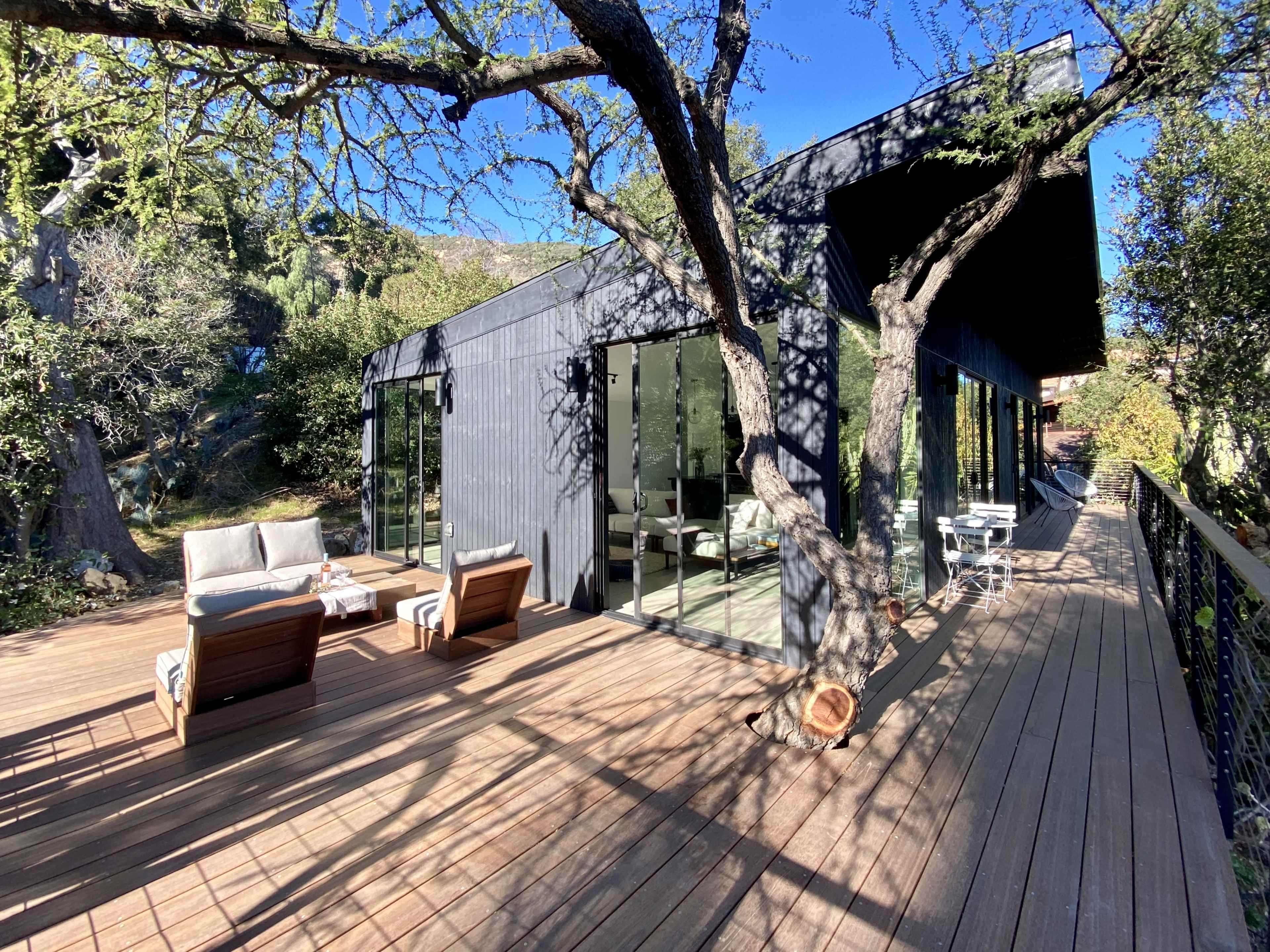 A modern black wooden house is surrounded by trees, featuring a spacious deck with seating arrangements.