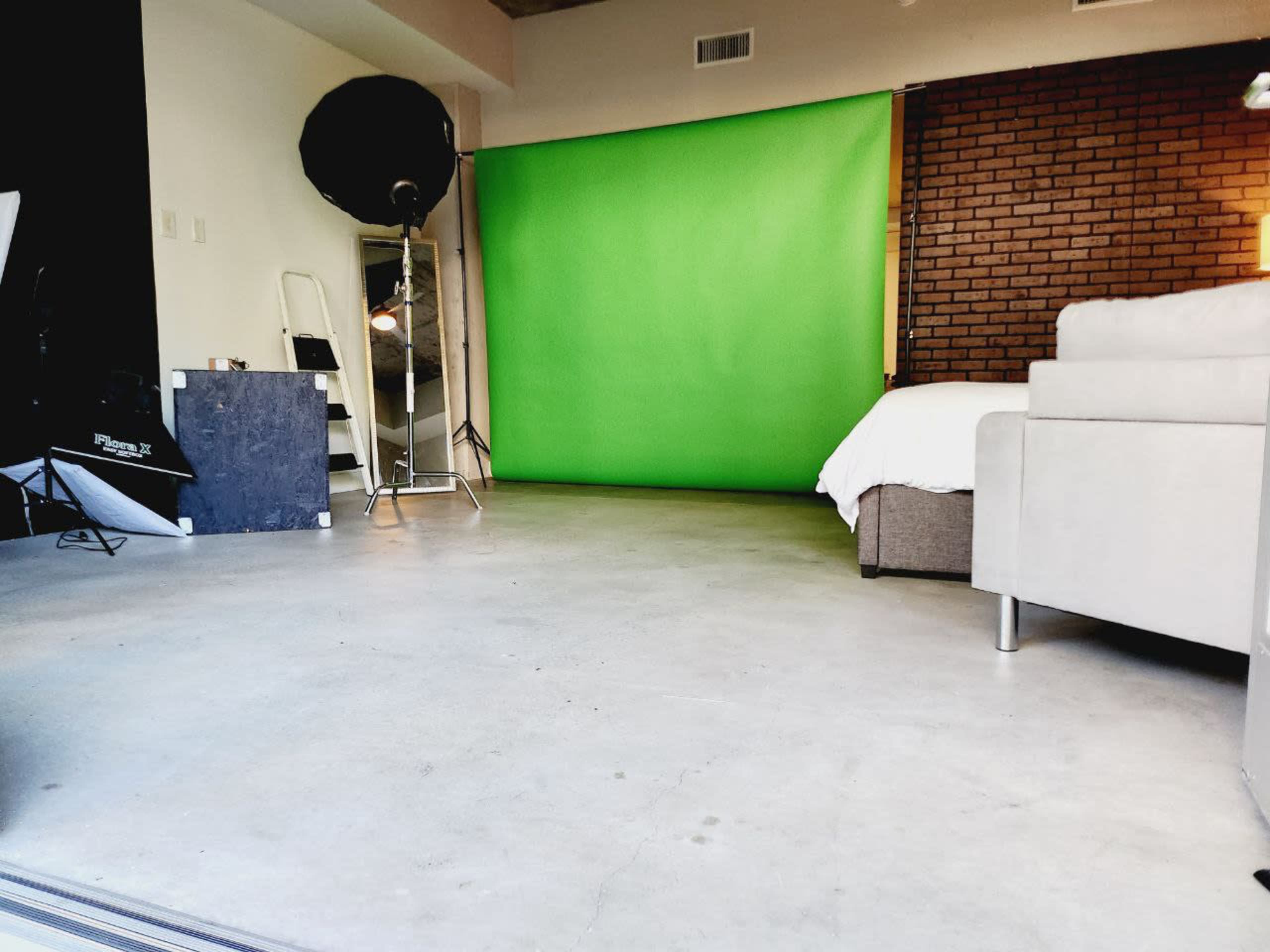The image shows a studio space featuring a green backdrop, a brown brick wall, a white sofa, and a few lighting equipment pieces.