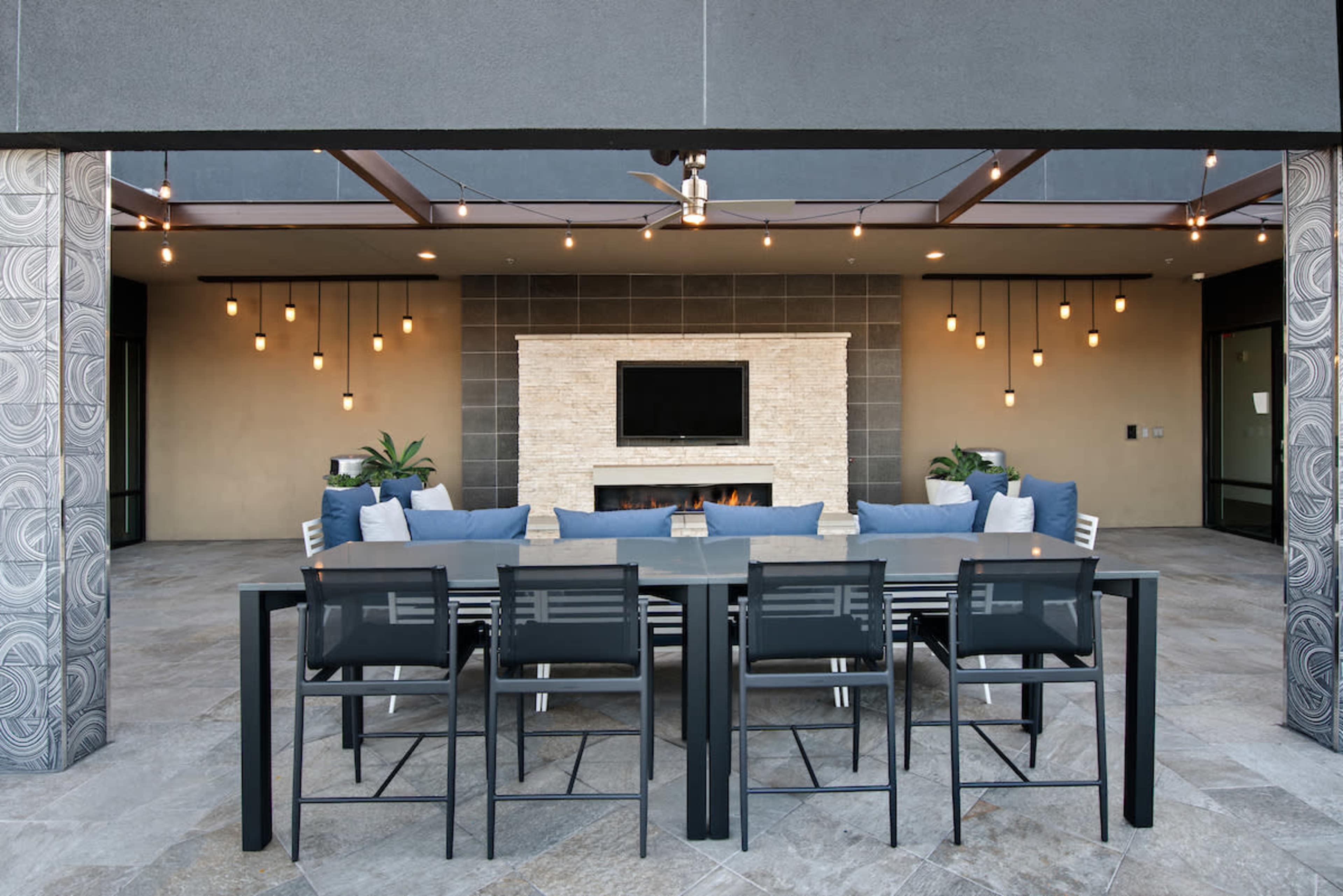 A modern outdoor patio with a dining table and seating area, featuring a stone fireplace and suspended light fixtures.