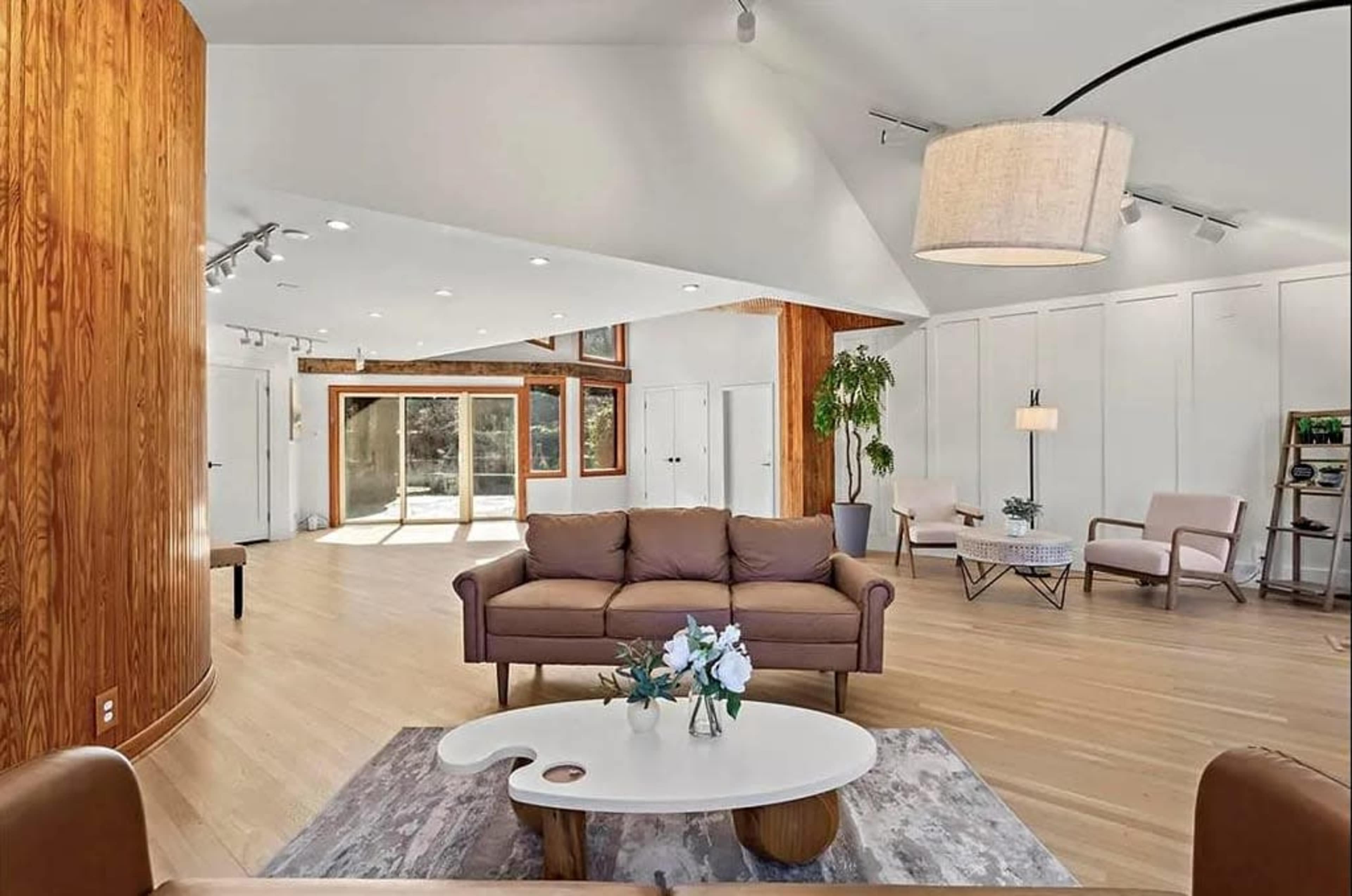 The image shows a spacious modern living room with a light wood floor, a brown sofa, a circular coffee table, and large windows allowing natural light to enter.
