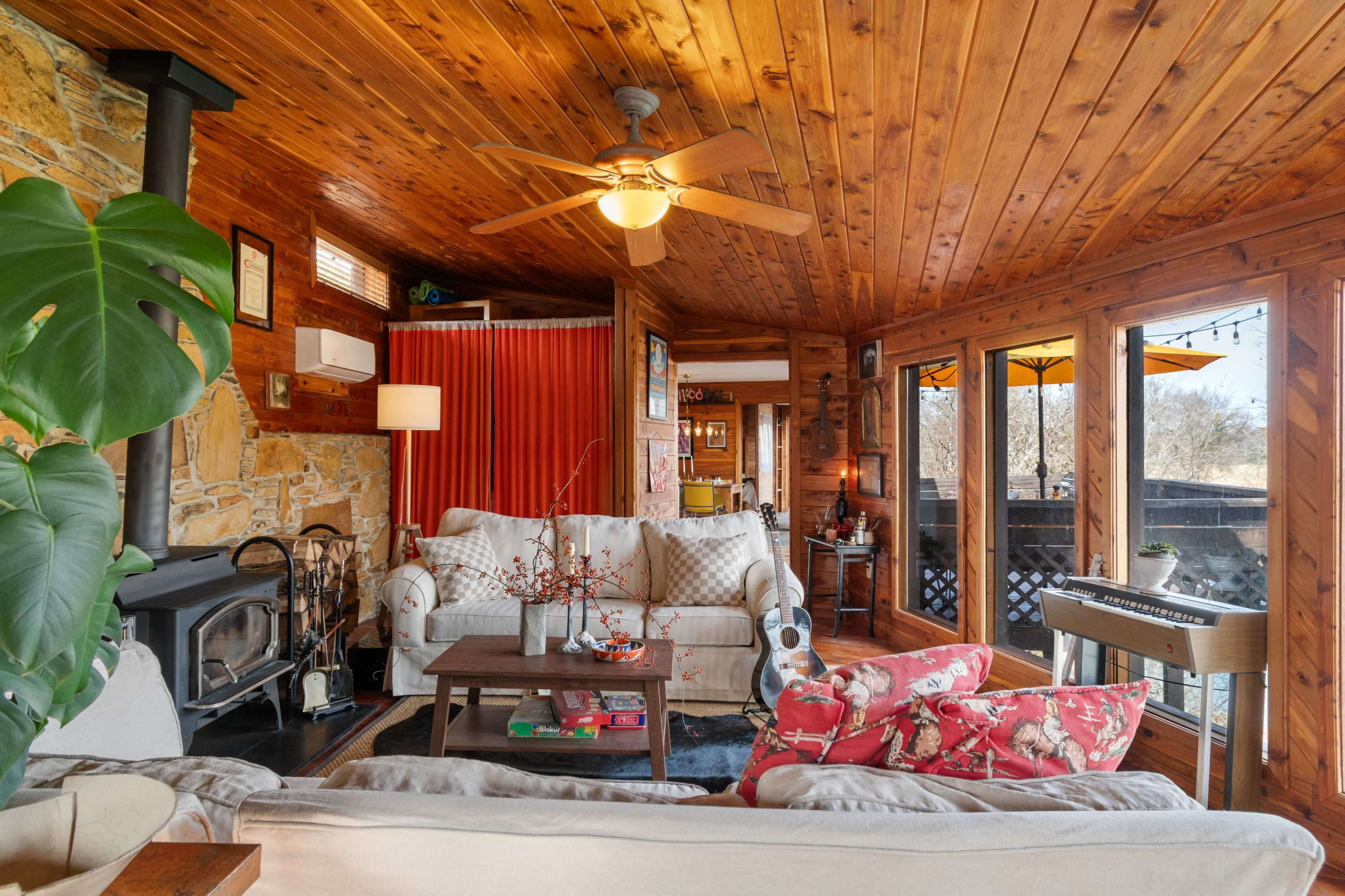 The image shows a cozy living room with wooden walls and ceiling, featuring a sofa, armchairs, a coffee table, a fireplace, and large windows leading to an outdoor area.