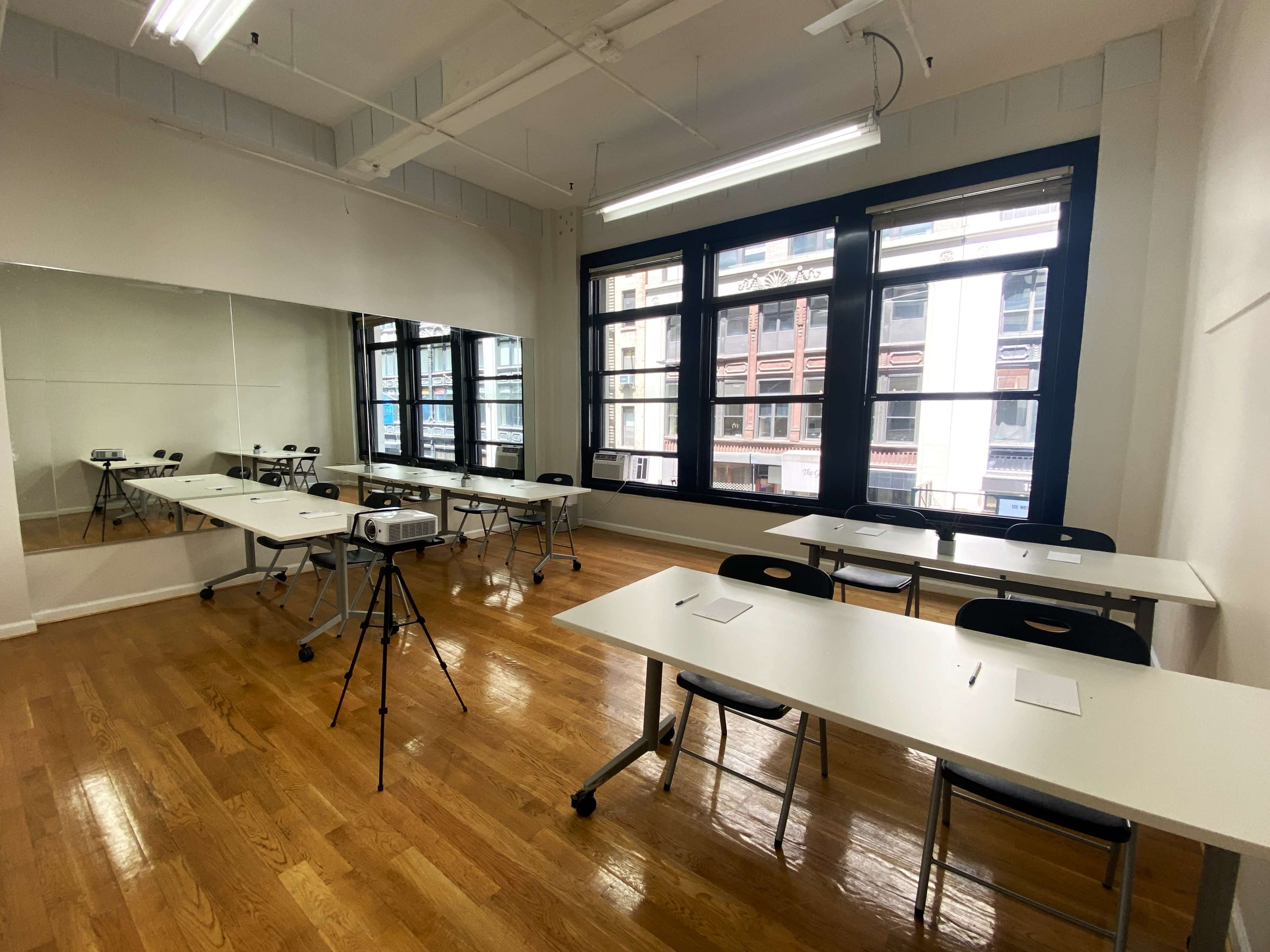 A well-lit classroom features several tables and chairs arranged around the space, with a projector on a tripod in front of a large mirror and windows.