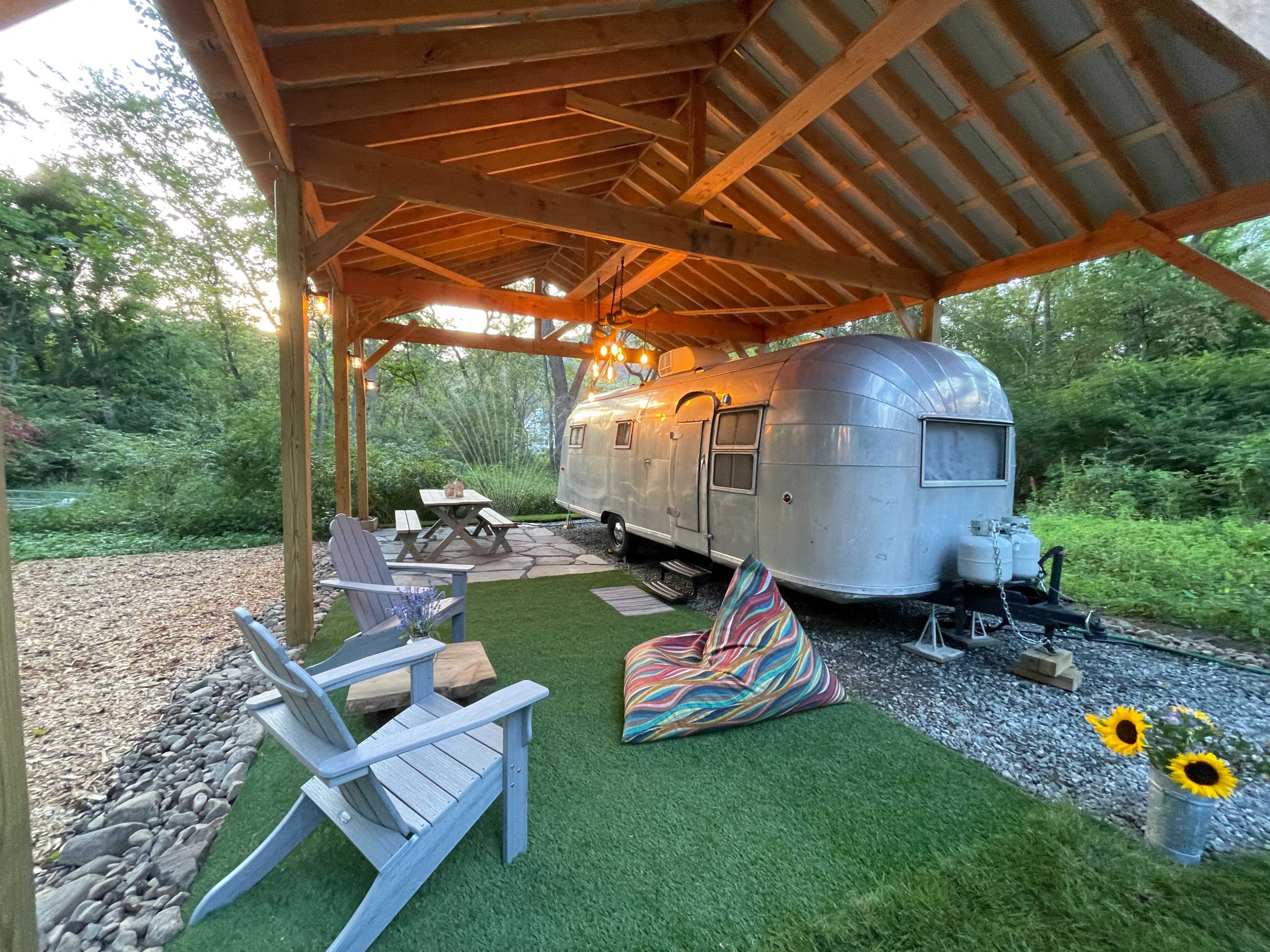 A silver trailer is parked under a wooden shelter with outdoor seating and a landscaped area featuring grass and decorative plants.