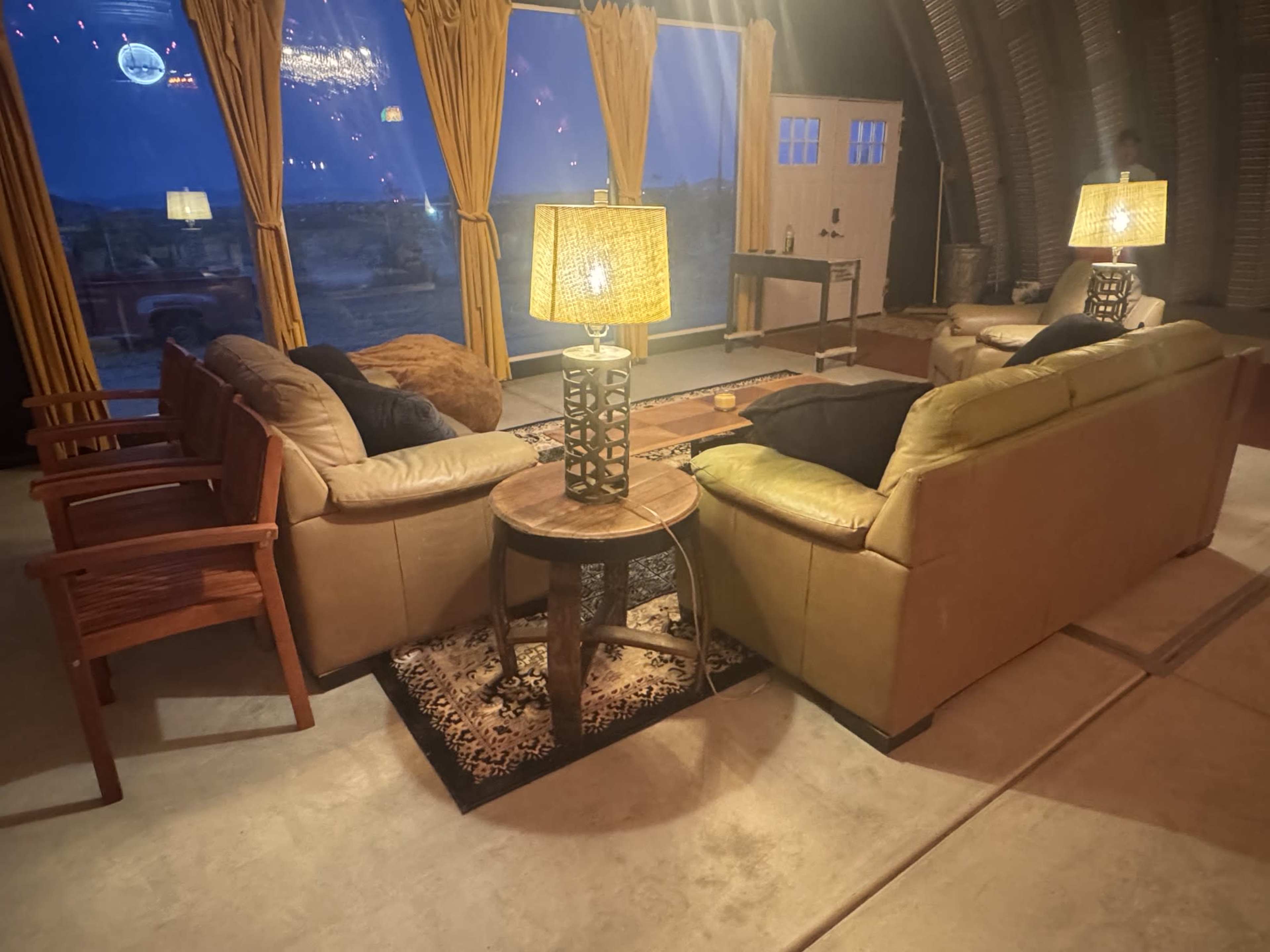 The image shows a cozy living space with two sofas, a wooden table, and lamps, set against large windows revealing an evening landscape.