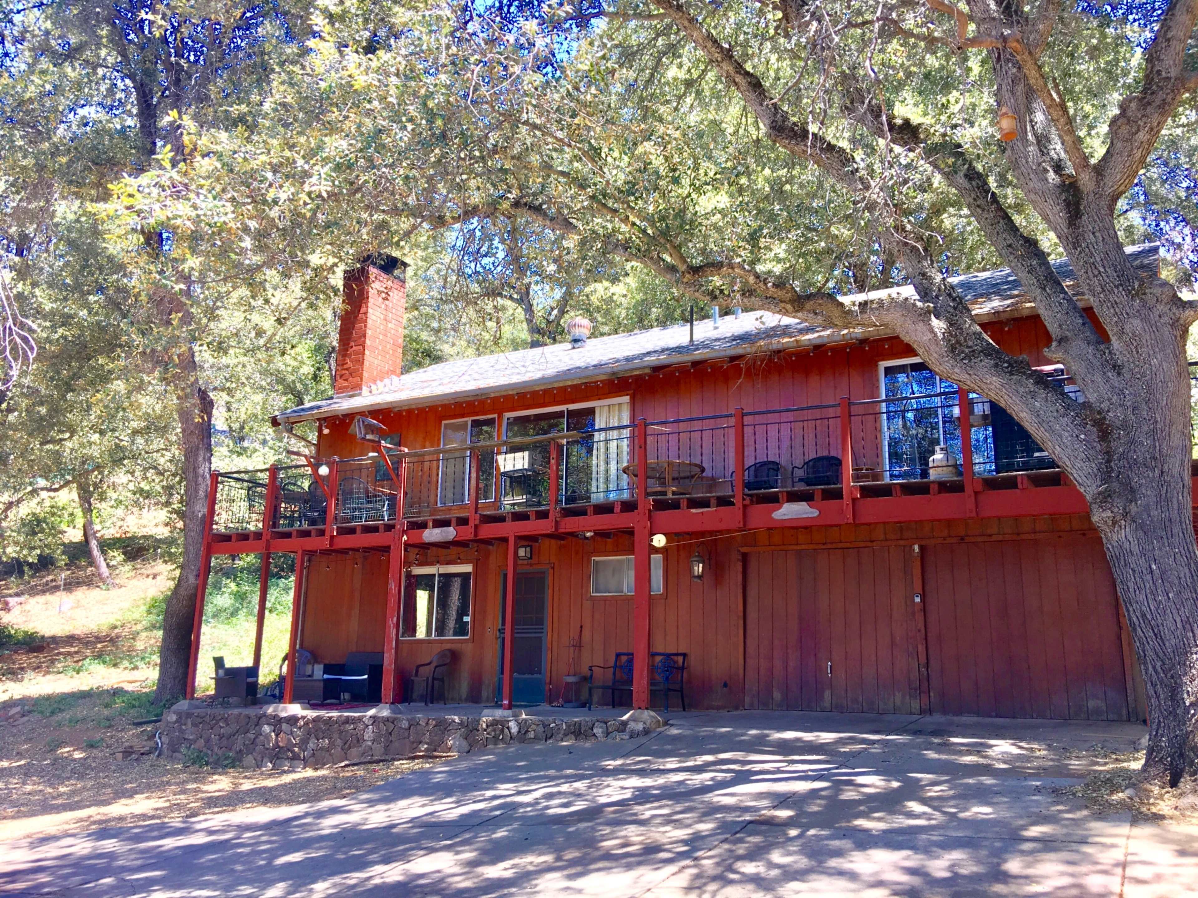 A two-story red wooden house with a balcony and stone accents is surrounded by trees.