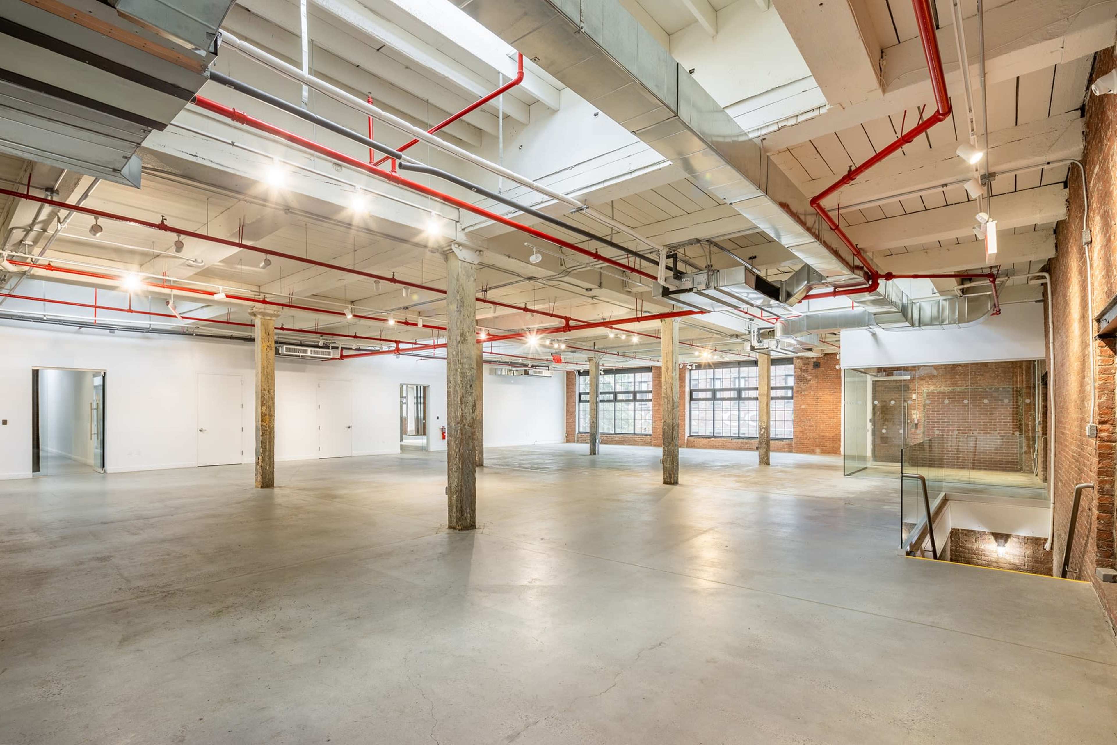 A spacious industrial-style room with concrete floors, exposed brick walls, and overhead ducts and pipes.
