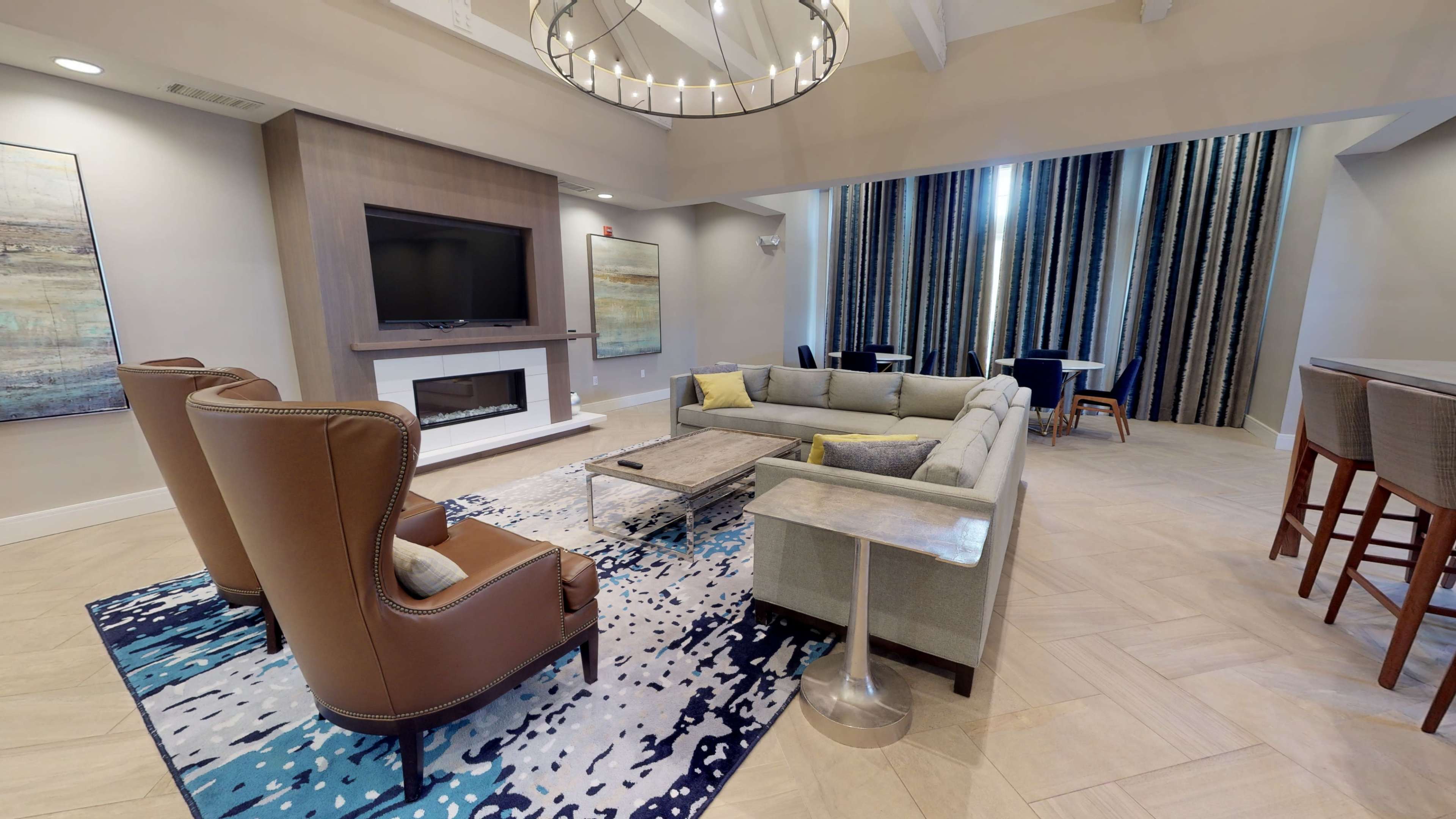 A modern lounge area featuring a sectional sofa, an armchair, a coffee table, a large flat-screen television, and a dining area in the background.